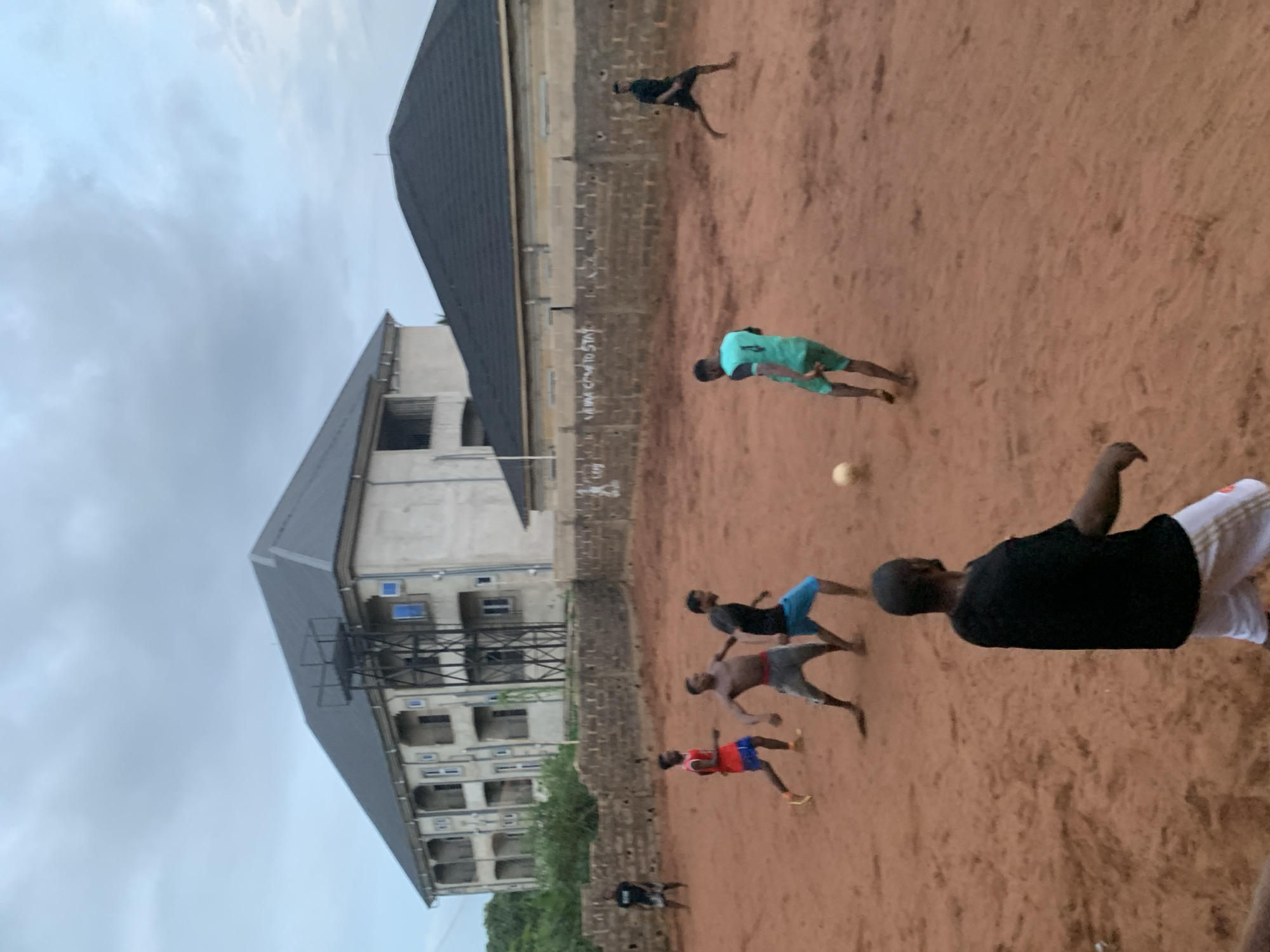 The image depicts a group of people playing soccer on a dirt field in Ukwu-Abwa, Nigeria. The scene is dominated by the reddish-brown dirt field, where a soccer ball is visible. Several individuals are actively engaged in playing; they are wearing casual clothing, including shorts and shirts. One individual is wearing a bright green jersey. In the background is a multi-story building with a dark roof, appearing to be partially under construction or in a state of disrepair. The sky is overcast. A section of a brick wall is also visible. The overall setting appears to be an undeveloped or poorer area, possibly on the outskirts of the city. The image is rotated counter-clockwise.