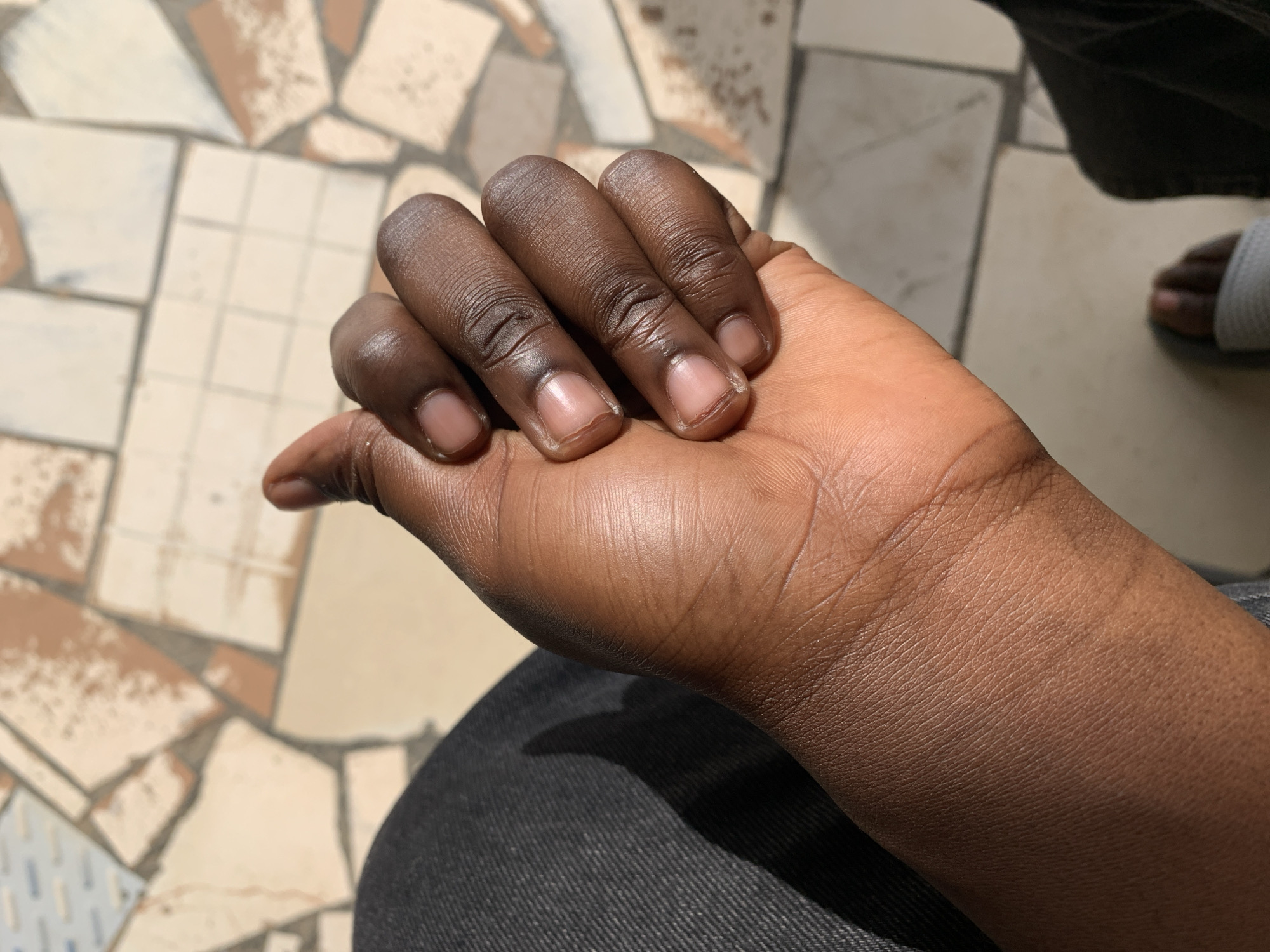 The image shows a close-up of a person's hand, with the fingers curled inwards and the thumb extended upwards. The skin is dark brown and appears smooth, with visible lines on the knuckles and palm. The fingernails are short and unpolished. The person is wearing dark gray denim pants, with a shadow cast across the leg. The background consists of a tiled floor with irregular shapes in shades of cream, beige, and brown, suggesting an outdoor setting. The lighting indicates it is daytime, with sunlight casting shadows. The overall scene is casual and appears to be a personal moment captured in a straightforward manner.