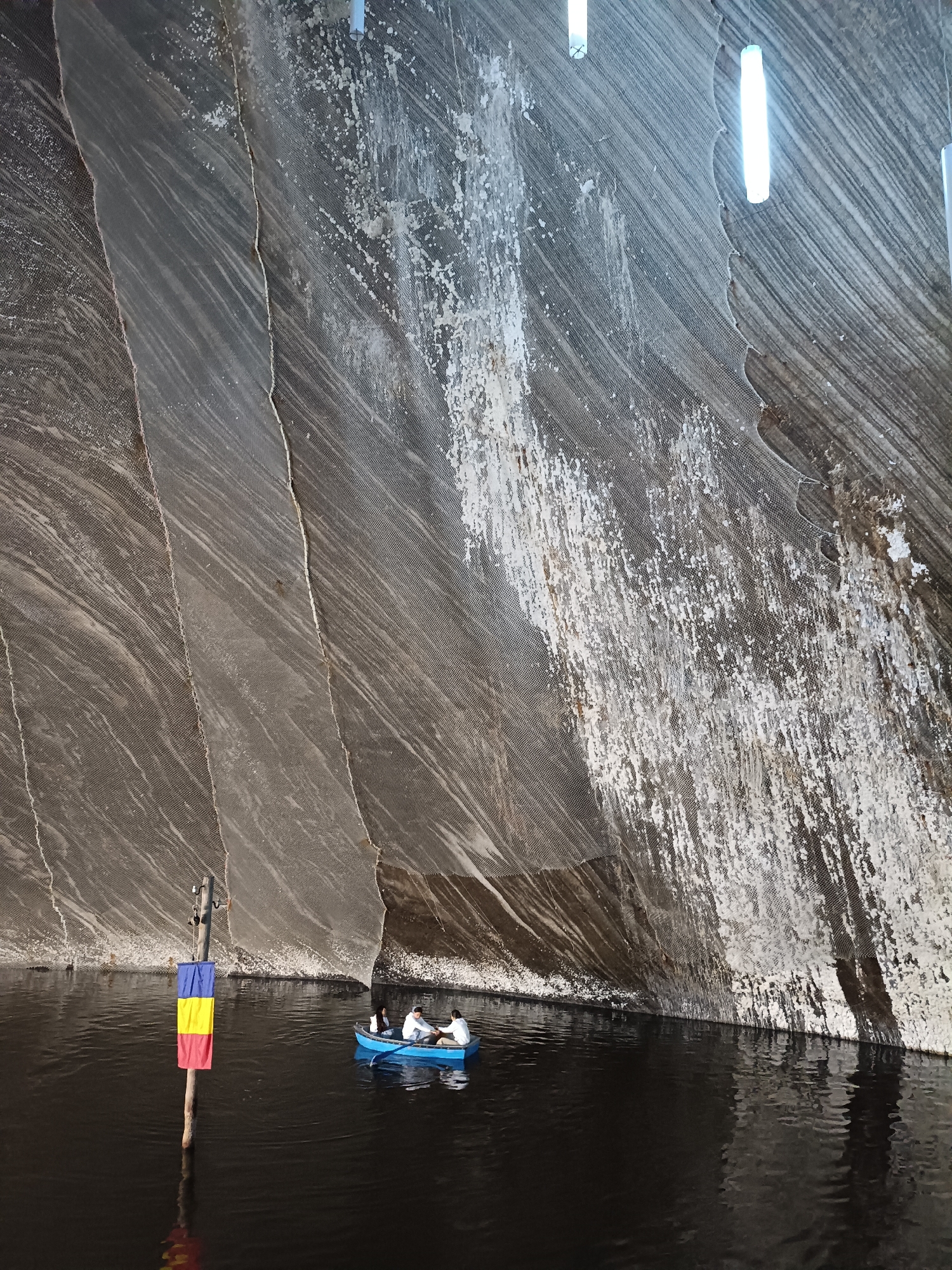 The image depicts an underground salt mine in Turda, Romania. A dark, still body of water fills the foreground, with a small blue boat carrying three people gently navigating the surface. The boat's occupants appear to be engaged in conversation or enjoying the experience. To the left, a wooden pole stands in the water, adorned with the Romanian flag, its colors of blue, yellow, and red clearly visible.
The background is dominated by the cavernous walls of the salt mine, characterized by dramatic, stripy rock formations and patches of white salt deposits. The sheer scale of the mine is emphasized by the low vantage point from the boat, making the walls appear immense and imposing. The lighting, provided by several long, cylindrical fixtures hanging from the ceiling, casts a cool, white glow, illuminating the textured surfaces of the salt and rock. The overall atmosphere is serene and awe-inspiring, highlighting the unique natural beauty and industrial history of the location. The scene appears to be set during daylight, though the underground environment creates a subdued illumination.