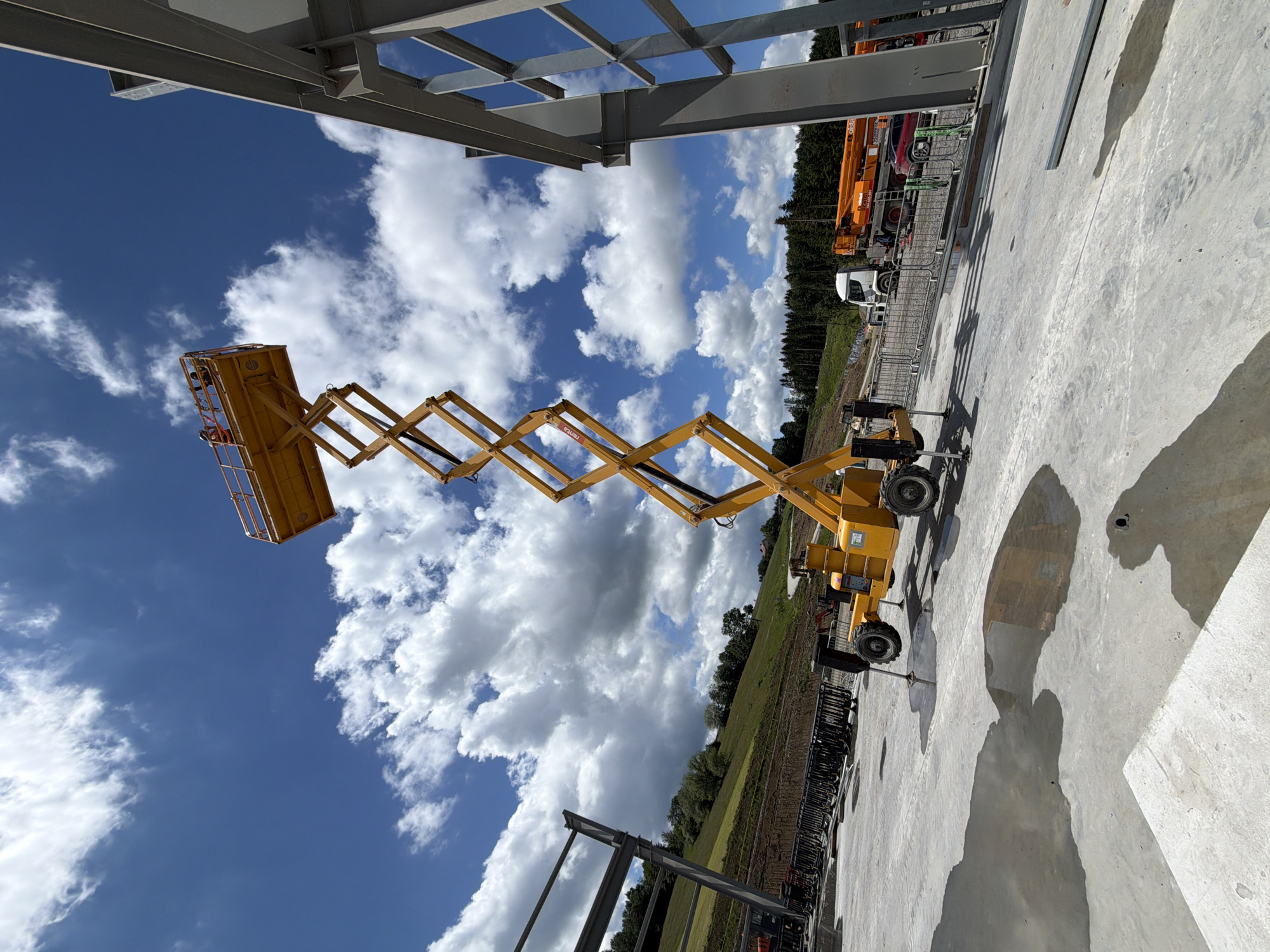 The image depicts a construction site scene under a partly cloudy sky. Dominating the center is a tall yellow scissor lift, extended upwards, with two individuals visible in the platform at the top. To the right, a section of a grey steel building frame is under construction. In the background, a hilly landscape covered with trees is visible, along with a white truck and an orange crane or similar heavy machinery. The foreground shows a wet concrete surface with puddles reflecting the surroundings. The overall impression is one of ongoing construction work in an outdoor, somewhat rural setting. The location is Janowice, Poland.