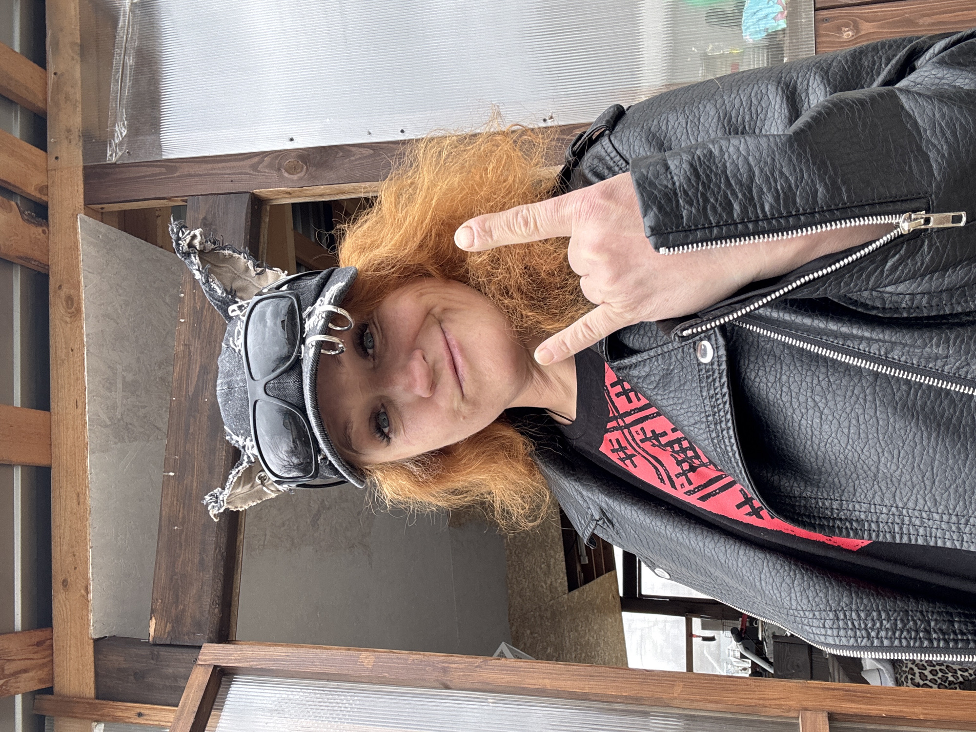 A woman with orange hair is posing for a photo. She is wearing a black leather jacket over a black t-shirt with a red design, and a grey cap with attached sunglasses and decorative elements resembling animal ears. She is making a "rock on" hand gesture. The setting appears to be a semi-enclosed wooden structure, possibly a shed or porch. There are wooden beams and what seems to be corrugated plastic sheeting in the background. The location is Chaikovsky, Russia.