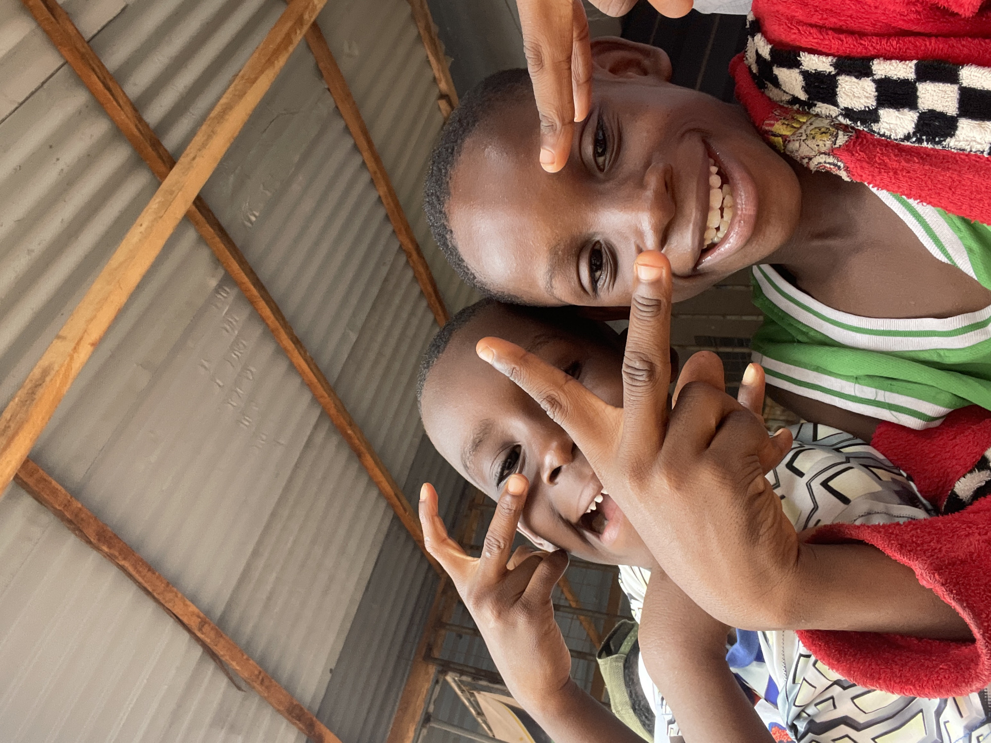 Two dark-skinned children pose for a selfie indoors, making a peace sign with their hands. The child on the top is wearing a green shirt with white stripes at the neckline, as well as a red towel with black and white checkered trim. The child on the bottom wears a patterned white shirt. Behind the children is a roof made of corrugated metal attached to wooden beams. The setting is Onitsha, Nigeria.
