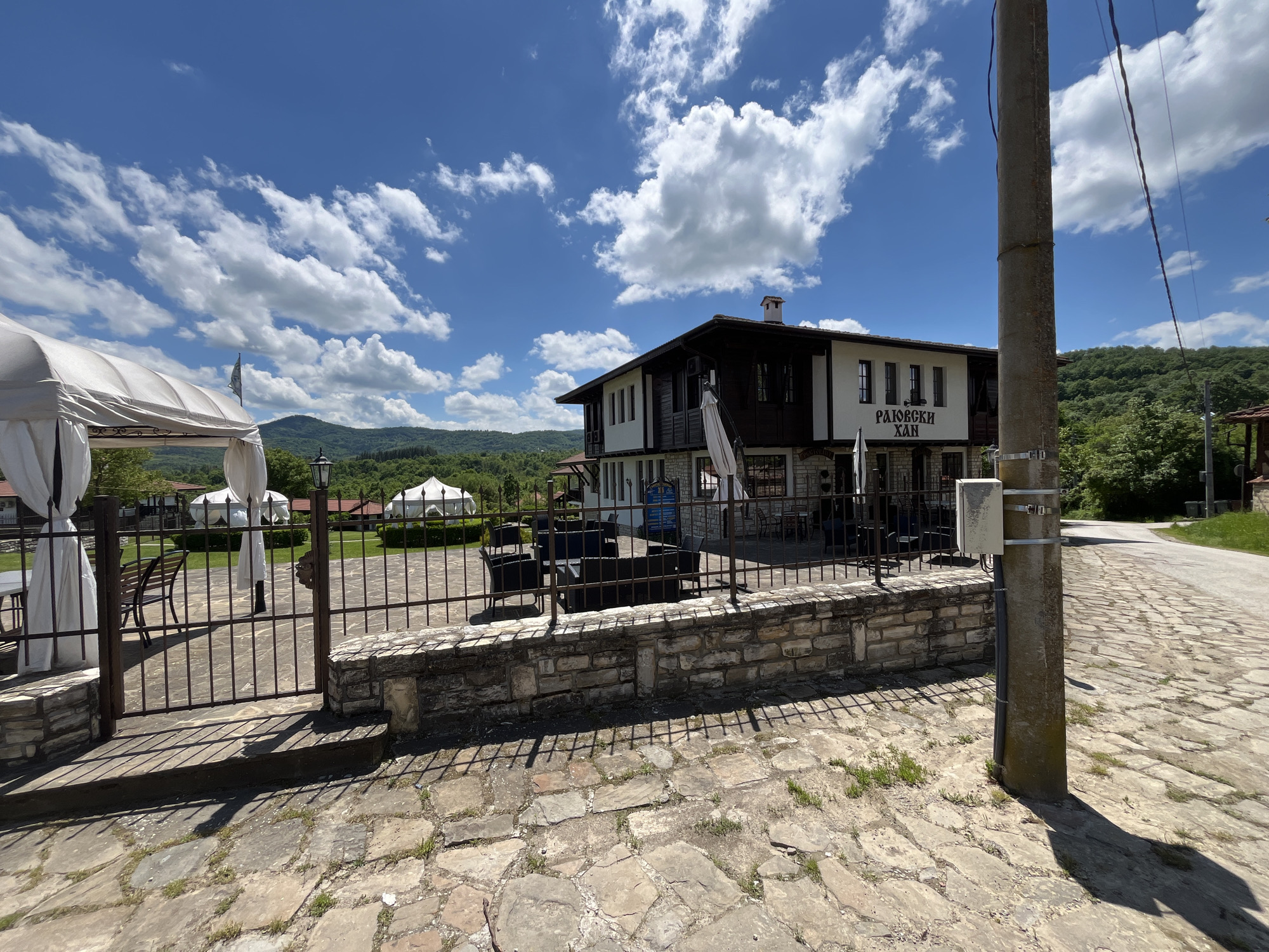 The image shows the exterior of a two-story building with the sign "РАЮВСКИ ХАН" (Rayuvski Han) in Rayuvtsi, Bulgaria. The building has a white facade with dark brown wooden accents and a stone foundation. In front of the building is a stone wall with a metal fence. Outdoor furniture, including tables and chairs, are visible behind the fence.
To the left, there is a white tent-like structure with draped curtains. The background features rolling green hills and a bright blue sky with scattered white clouds. In the foreground on the right, there is a concrete utility pole with a junction box attached. The ground is covered with stone paving.