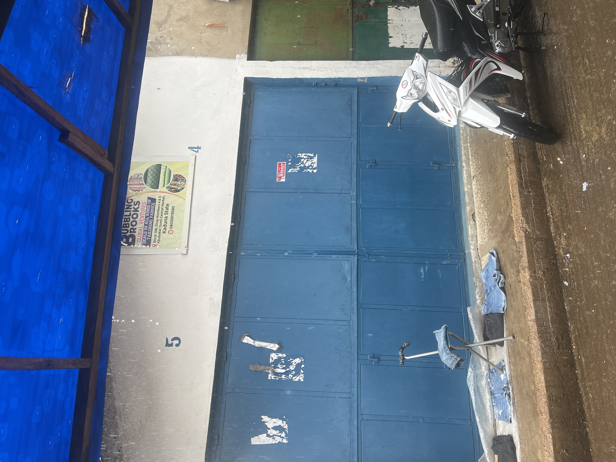The image depicts an outdoor scene in front of a building with blue metal doors. A white and red motorcycle is parked to the right. In front of the doors, several pairs of denim shorts are laid out on the ground and on a folding stool, which is also draped with denim. To the left, a black object, possibly a bag, is also on the ground. The building's facade is white with blue numbers "5" and "4" painted on it. A sign with "BUBBLING BROOKS GLOBAL VENTURES - DEALS IN ALL KINDS OF TAILORING FABRICS" is visible. The environment appears wet, with raindrops falling and puddles on the ground, suggesting it is raining. The scene suggests a commercial or market setting, possibly a tailor's shop or fabric stall.