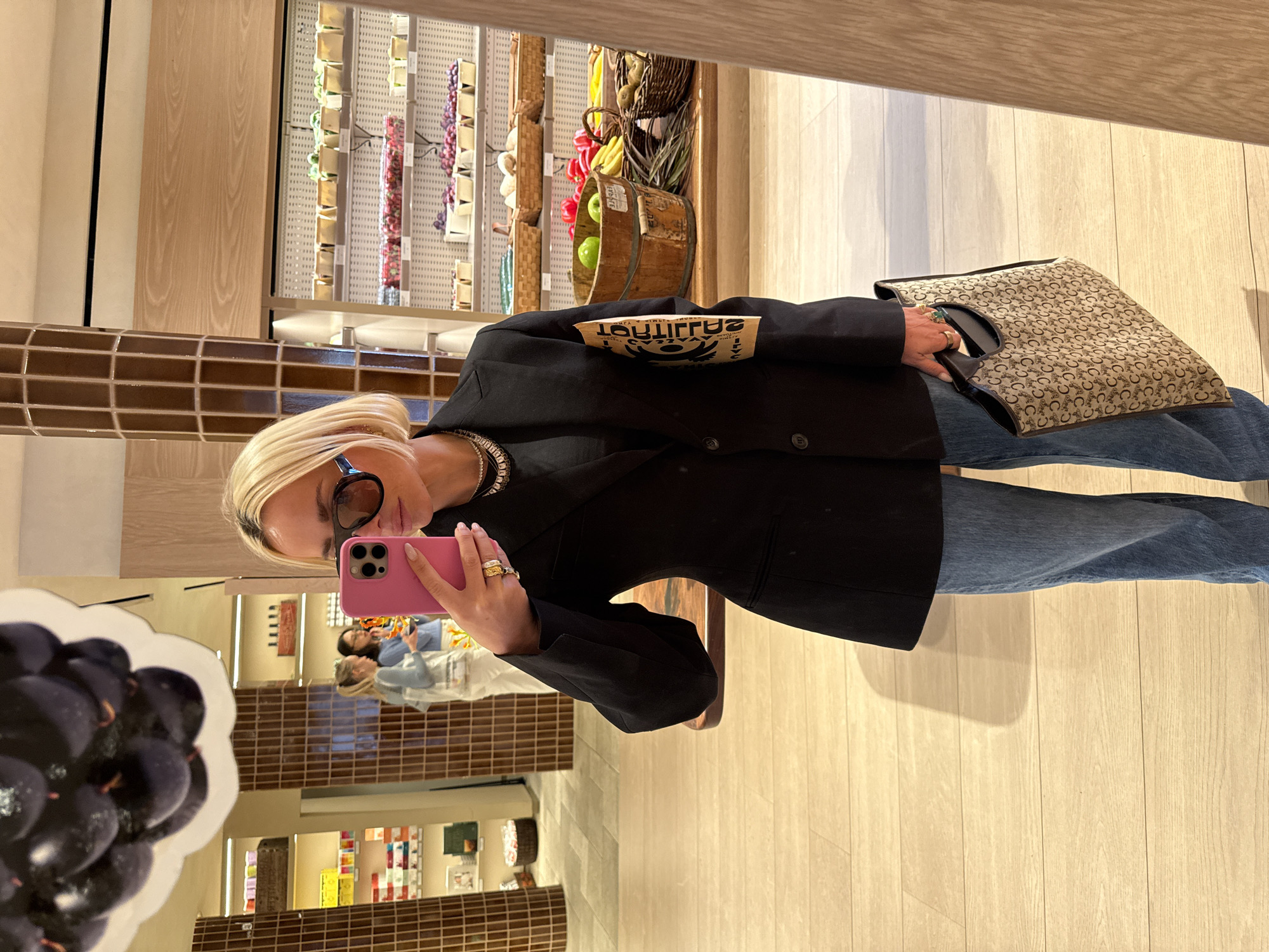 The image is a selfie taken by a person with blonde hair, wearing a black blazer, blue jeans, and holding a pink iPhone in front of a mirror. The reflection shows shelves with various items on display, a person in a light-colored outfit in the background, and a decorative piece resembling a bunch of grapes. Based on the products on display and the overall interior design, it is likely that the location is Erewhon Market in the city of New York, United States of America.