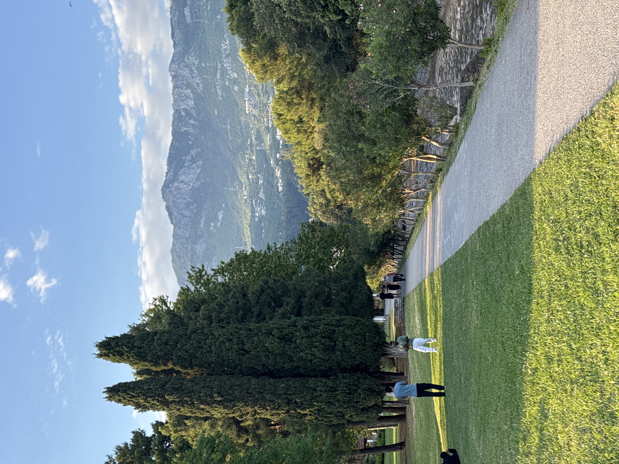 A long, horizontal, eye-level shot depicts a gravel path bordered by a green lawn and various trees, with a mountain range in the distance. The sky is partially visible in the upper left, displaying a blue sky with scattered clouds.
The gravel path, situated to the right, extends into the distance. A stone wall runs parallel to the path, separating it from a wooded area with lush green trees.
To the left, a well-maintained lawn occupies a significant portion of the frame. Several people are present on the lawn. One individual is wearing a blue shirt and black pants. Another person, in white clothing, appears to be lying down or bending over. Further down the path, other figures are visible.
In the background, a rugged mountain range dominates the horizon. Small buildings or structures are visible on the mountainside, suggesting a settlement.
The overall scene evokes a sense of tranquility and natural beauty. It is possible that this is near Budva, Montenegro, as this area is known for its mountains and coastal scenery.