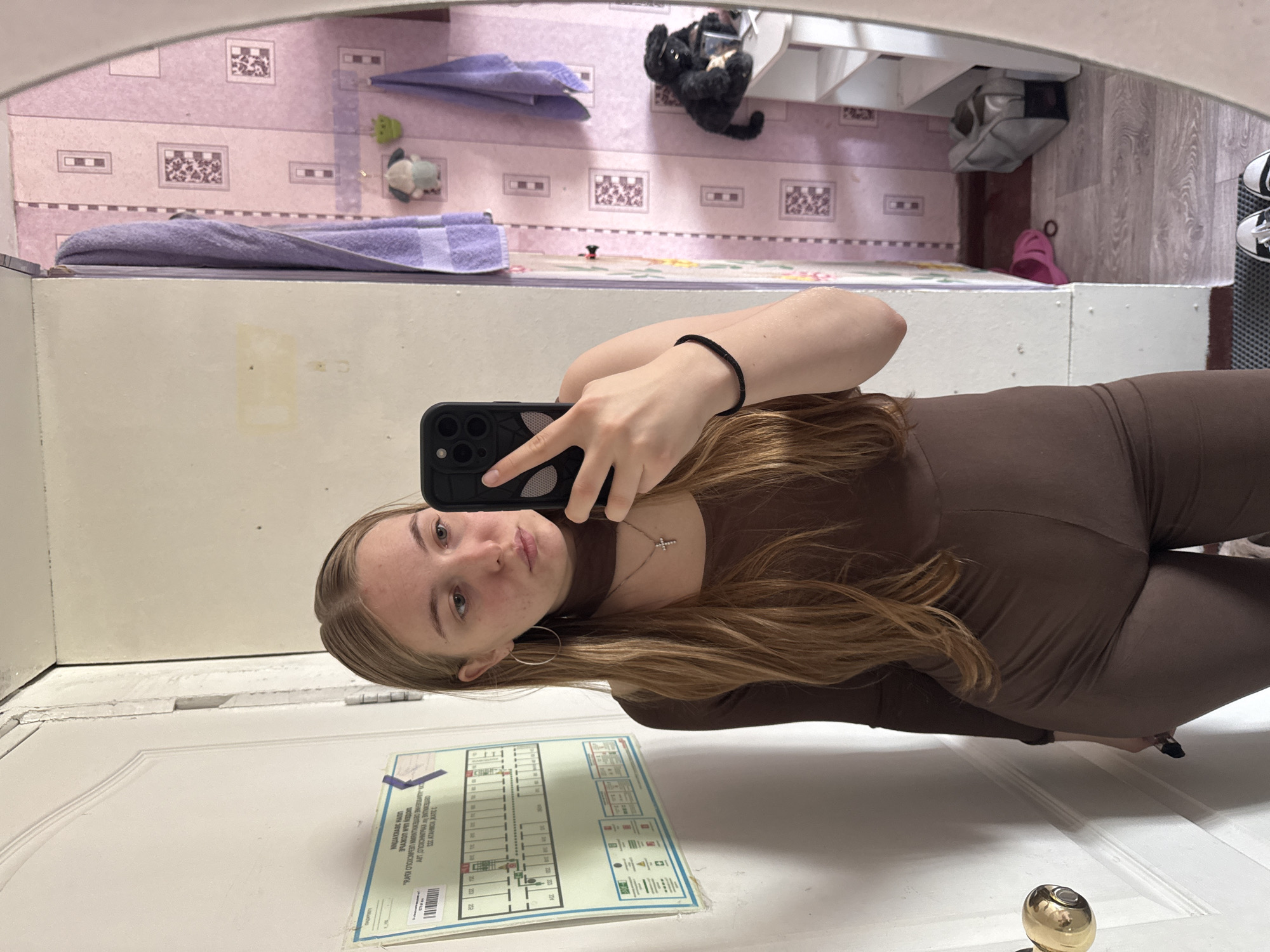 A young woman with blonde hair is taking a mirror selfie in what appears to be a bathroom or small room. She is wearing a brown long-sleeved top and matching leggings, with a silver cross necklace and hoop earrings. Her phone is black with a spider-web design on the case. In the background, a purple towel hangs on a rack, and the walls are covered in pink wallpaper with a floral pattern. A shelf unit is visible in the reflection, with a black stuffed animal on it. The room is dimly lit, and the overall impression is of a casual, personal space. Based on the wallpaper and overall aesthetic, the location is likely a residential interior in Perm, Russia.