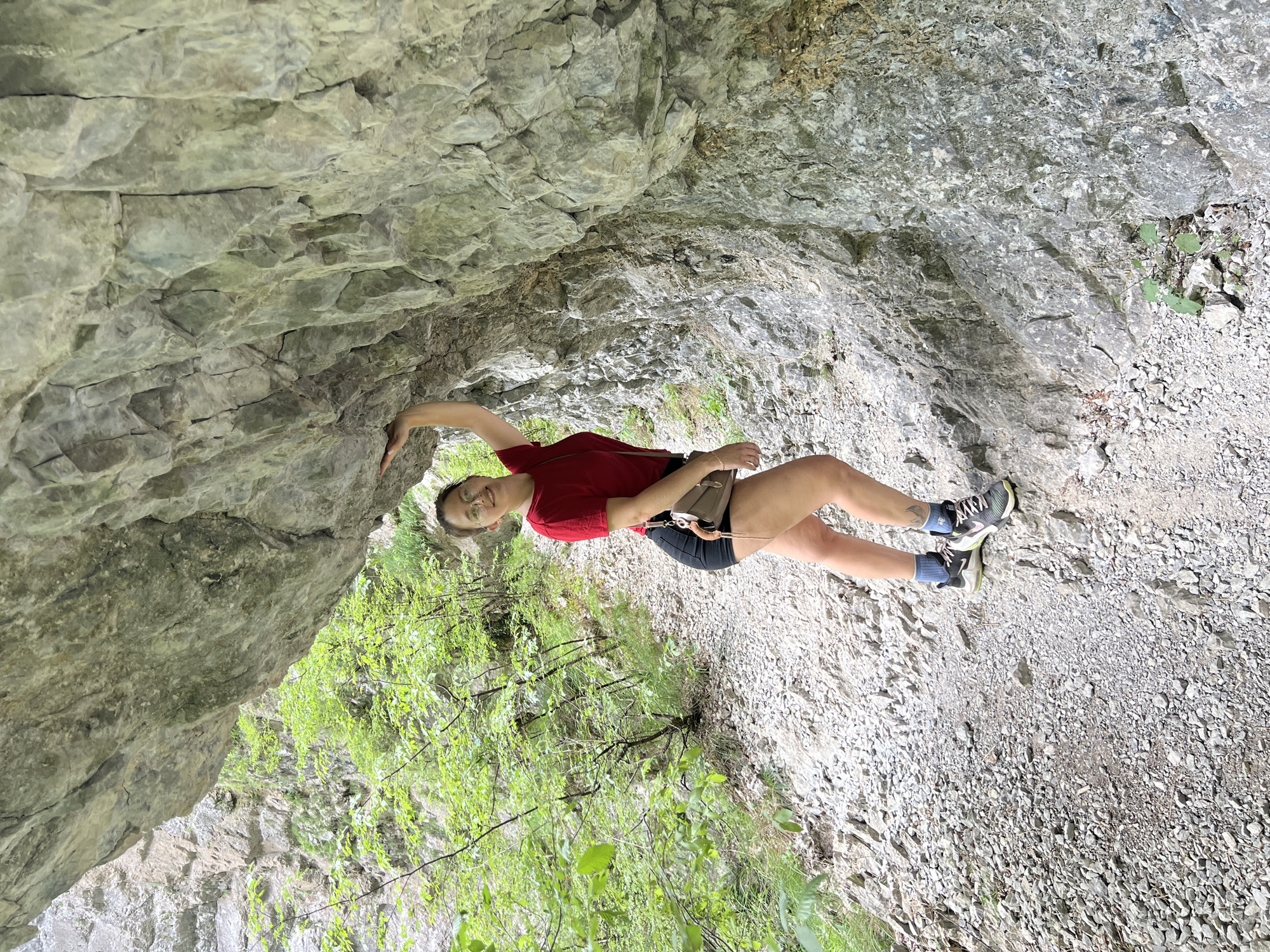 A woman with fair skin, wearing a red t-shirt, dark shorts, blue socks, and athletic shoes, is positioned against a large rock formation, potentially in a natural setting. She has a small bag hanging on her left hip. Her left hand is touching the rock face. The background includes the rock formation and some greenery. The scene appears to be outdoor in daylight. The location might be a hiking area. The image is oriented on its side, where the woman appears to be climbing the rock formation. She is smiling and looking towards the camera. Based on the prompt, the location is Lazba, Montenegro.