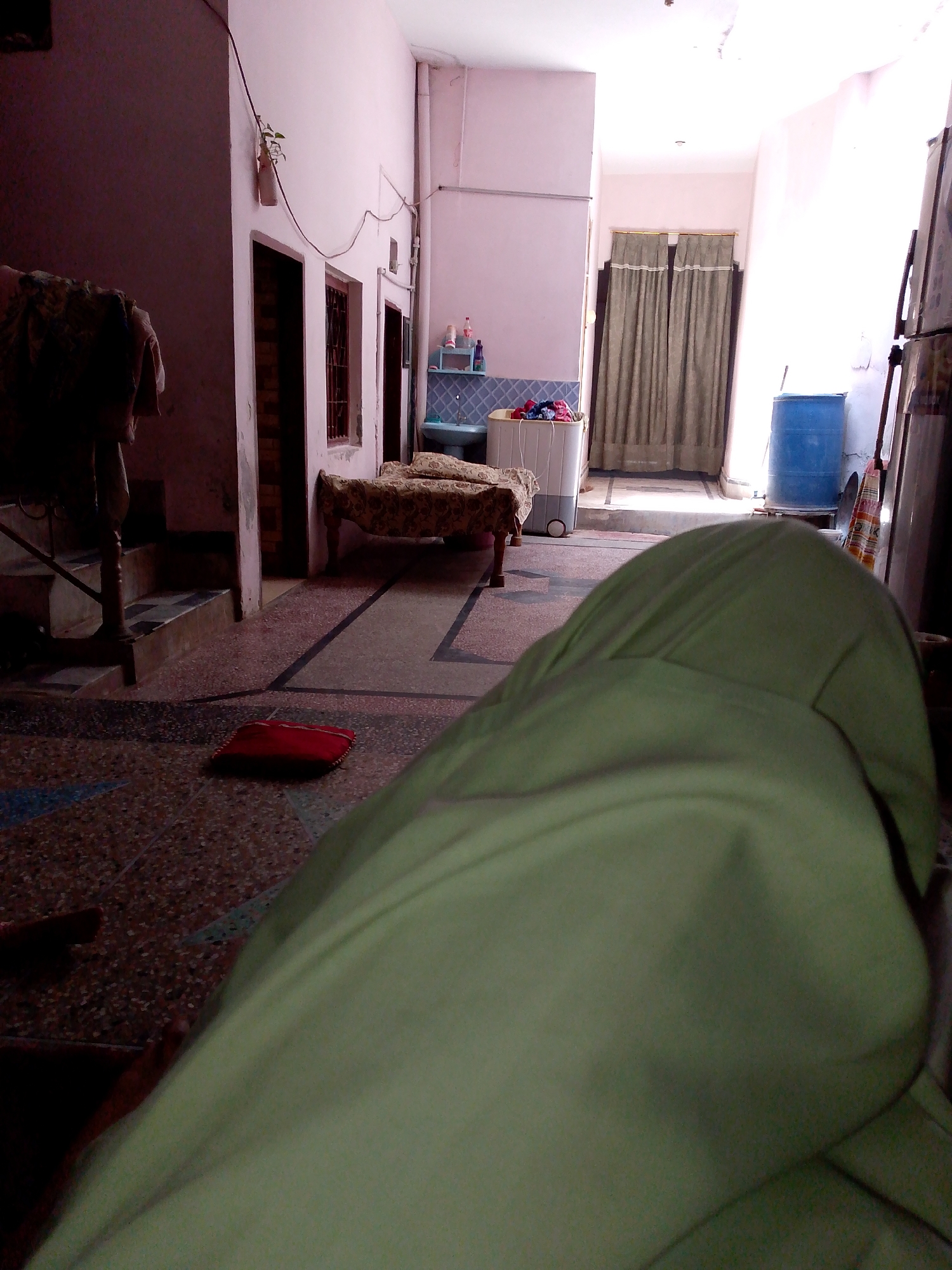 The image depicts an indoor scene of what appears to be a humble dwelling. In the foreground, a person's leg, covered in a light green fabric, is prominently featured, occupying a significant portion of the frame and suggesting a low-angle perspective. The flooring is a patterned mosaic of dark and light tiles, with a dark red cushion lying on it to the left. In the mid-ground, a narrow bed with a patterned bedspread is visible, positioned against a pink wall. To the right of the bed, a white top-loading washing machine is present, containing a load of clothes. Further into the background, a blue plastic barrel stands near a doorway draped with light brown curtains. The walls are painted a pale pink, and the ceiling is visible, appearing to be illuminated by natural light filtering in from the right side of the frame, possibly from a window or opening that is not fully visible. The overall environment suggests a lived-in space, possibly in a low-income area. The scene is devoid of explicit details that would pinpoint a specific city or country, making the location "Unknown, Unknown" appropriate.