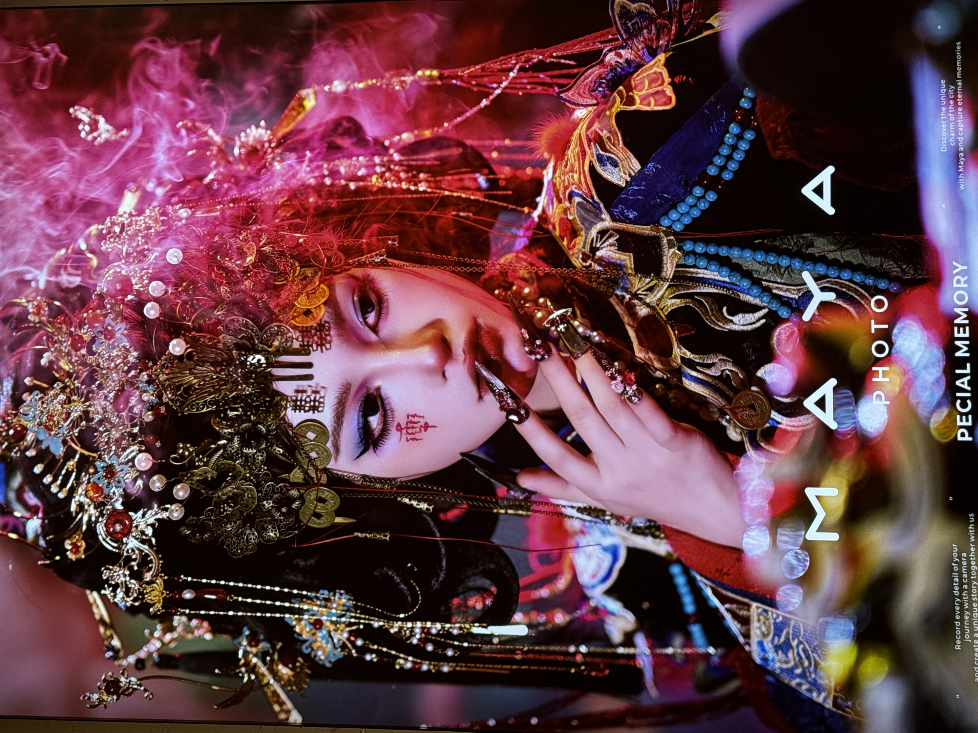 The image is a portrait of a young East Asian woman adorned in elaborate traditional Chinese attire and a headdress, which appears to be a historical or ceremonial costume. She has dramatic makeup, including bright blue eyeshadow and red facial markings on her cheek. Her fingernails are long and decorated with rhinestones. Her right hand is raised to her face, with her fingers touching her lips. The background is filled with swirling pink and red smoke, suggesting a dramatic or artistic setting. At the bottom of the image, there is text that reads "MAYA PHOTO" and "PECIAL MEMORY," along with smaller text about capturing memories. There is no discernible information about Yuzhong District, China, in the image itself.