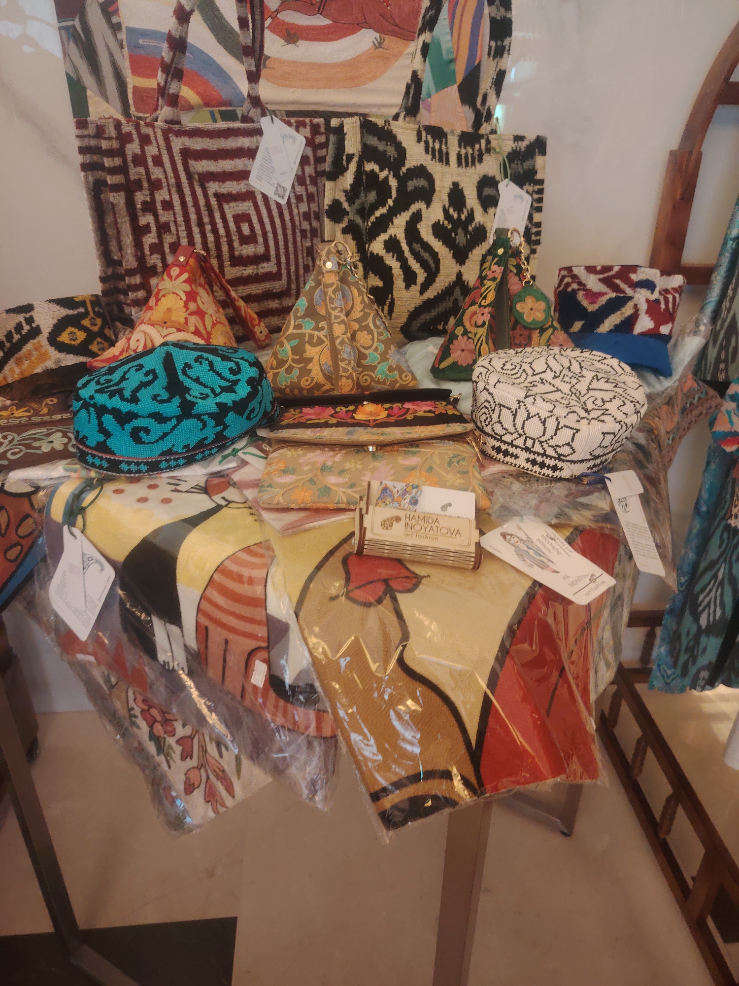 The image shows a display of traditional Uzbek textiles and accessories in a shop. The items include woven bags with geometric patterns, triangular embroidered pouches, and embroidered caps. One cap is turquoise with black designs, and another is white with black intricate patterns. There are also several flat, rectangular textile pieces with colorful, stylized figures and floral motifs, some of which are wrapped in plastic. A small wooden stand holds business cards with the name "HAMIDA INOYATOVA" and "ART FASHION." The display is set up on a table, and in the background, other textile items and a wooden rack are visible. The setting appears to be a shop in Konigil, Uzbekistan, a village known for its traditional textile production.