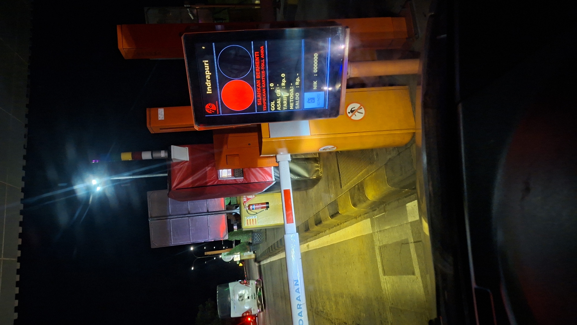 The image captures a nighttime scene at a toll booth, specifically identified as being in Sihomlhok, Indonesia. The foreground shows the interior of a vehicle, looking out through the windshield. A white toll barrier is lowered, blocking the path. To the right of the barrier is an orange toll booth structure with a digital display screen. The screen shows the toll plaza name "Indrapuri" and instructions in Indonesian, indicating to "PLEASE STOP" and "TAP YOUR E-TOLL CARD." It also displays financial information: GOL : 0, ASAL GB : -, TARIF : Rp.0, METODE : -, SALDO : Rp. -, and NIK : 000000. There are also icons of a camera and a person with a prohibition sign on the booth.
In the background, a white bus is stopped at the toll lane. Further back, streetlights illuminate the area, and the night sky is visible. The overall environment suggests a typical highway toll plaza operating at night.