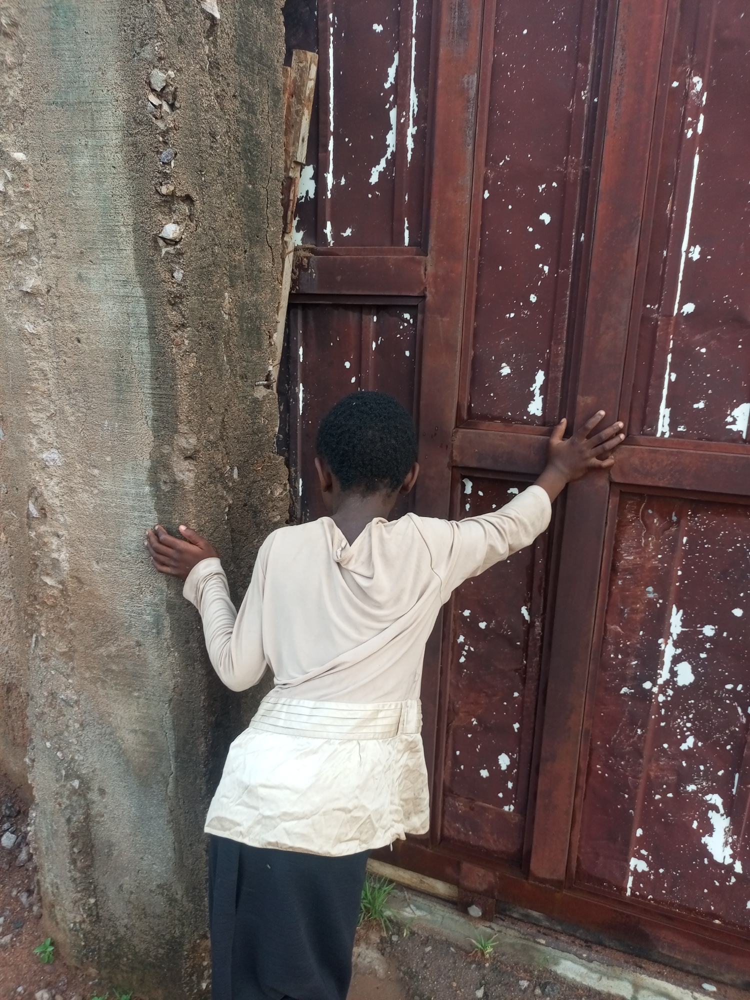 A young person, seen from the back, stands with their arms resting against a rough concrete wall on the left and a weathered, red metal gate on the right. The person wears a light-colored, long-sleeved shirt with a ruffled texture around the waist, over a dark skirt. Their hands are placed flat against the wall and the gate, suggesting they are leaning or holding themselves up. The concrete wall appears old and textured, with some small stones embedded in it. The metal gate is marked with white paint chips and shows signs of rust. The ground beneath is uneven and appears to be dirt with some sparse green vegetation. The scene seems to be an outdoor setting, possibly in a less maintained area of Tempure, Nigeria.