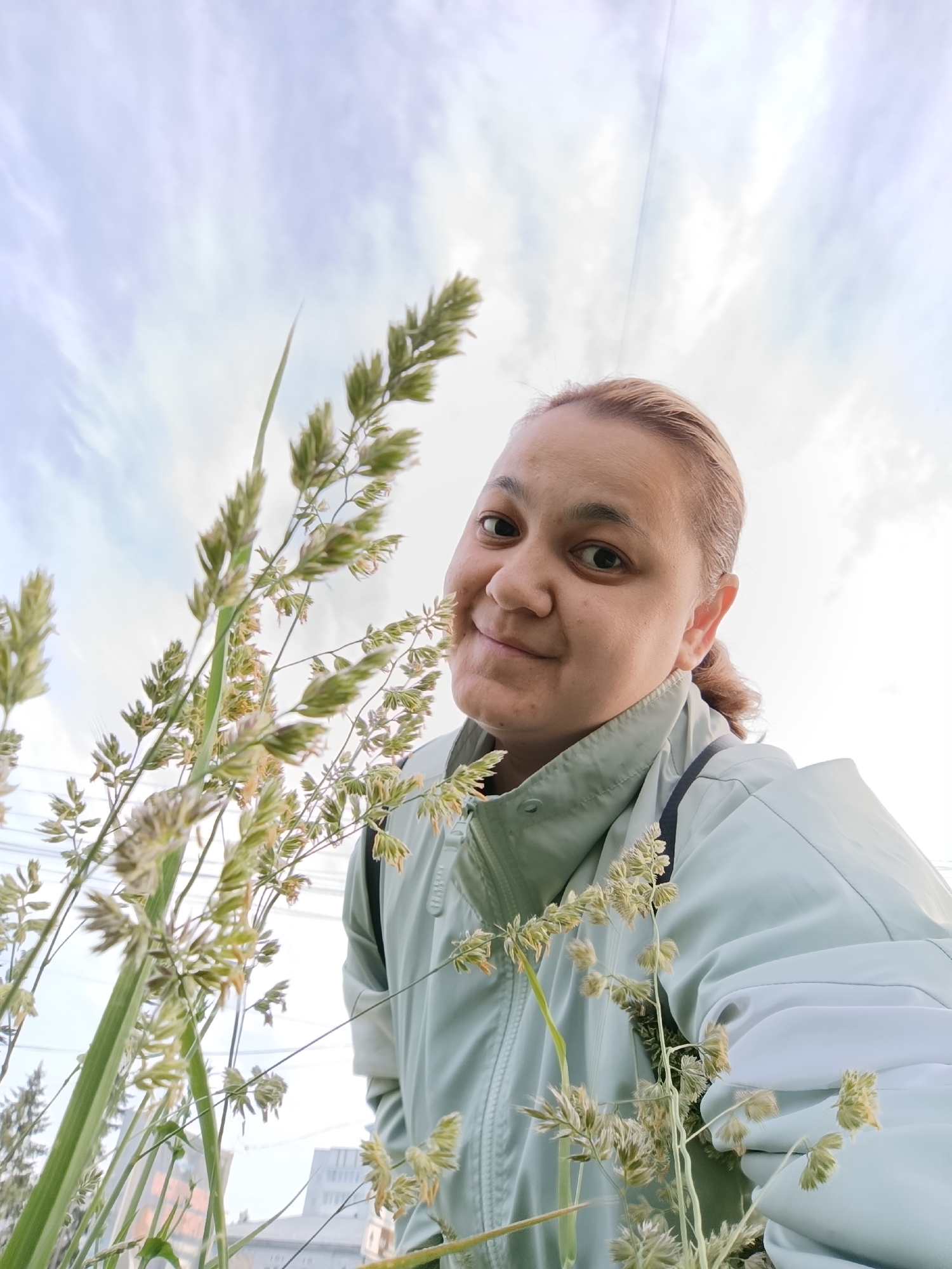 A low-angle portrait captures a woman with light brown hair, pulled back, smiling gently at the camera. She wears a light mint-green or pale blue jacket. The image is taken from below, looking up through a dense arrangement of tall, feathery grass that occupies much of the foreground and left side, framing her face on the upper right.
The sky, visible above and behind the grass, is overcast or partly cloudy with soft white and light gray clouds, indicating daytime with diffused light. In the distant background, behind the grass and beneath the sky, faint urban elements can be discerned, including the upper parts of a multi-story building and what appear to be utility lines or wires. A hint of greenery from distant trees is also visible. Faint, partially visible text or numbers, such as "161 78," can be seen on a structure in the background.
The setting appears to be an area where natural elements like tall grass meet an urban landscape, possibly a park, field, or roadside in Chaikovsky, Russia. The woman's expression conveys a calm and friendly mood.