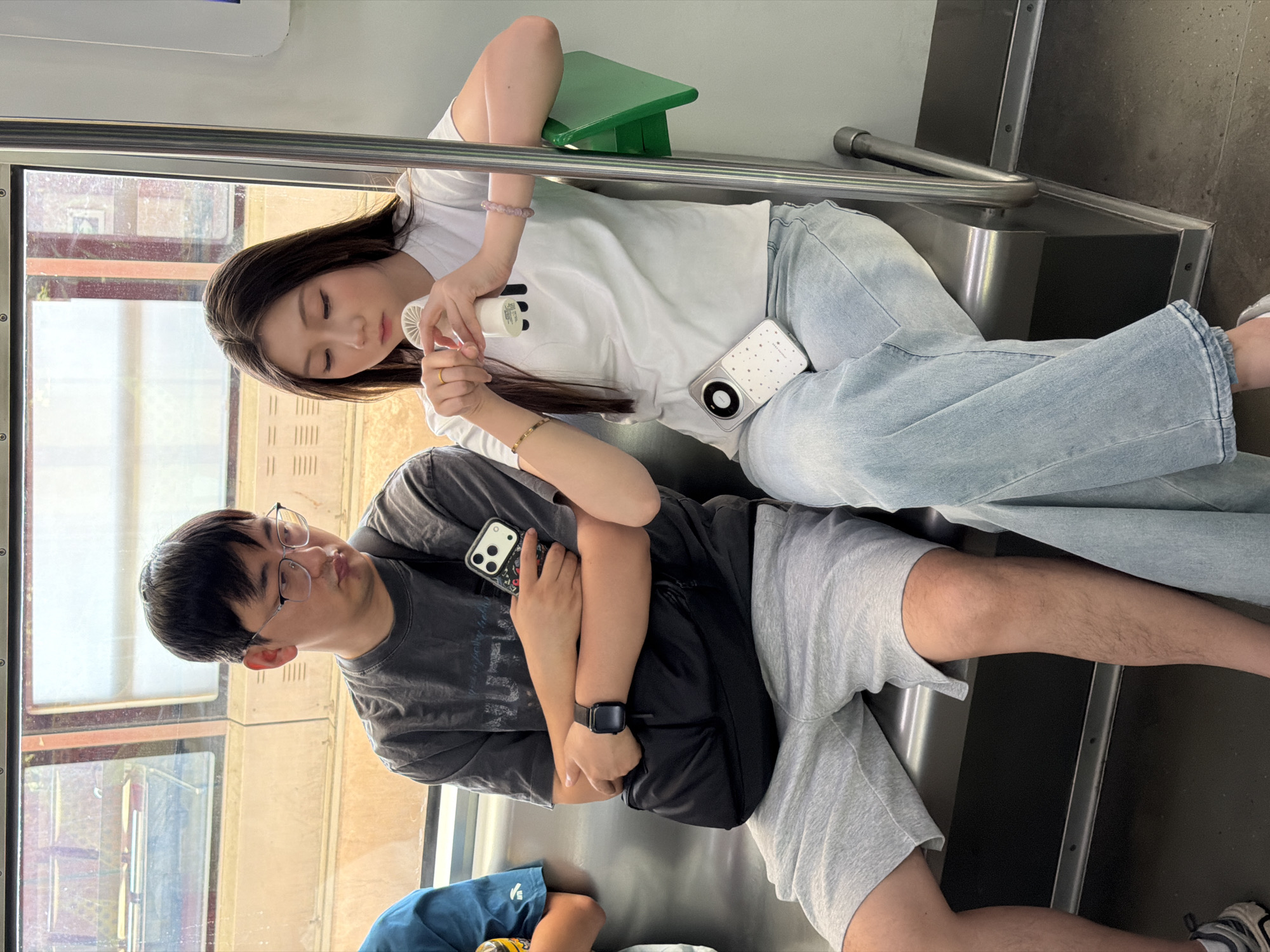 Two individuals are seated on a metal bench inside a public transit vehicle, likely a subway or light rail, in the Yuzhong District of China. The man on the left, wearing glasses, a dark grey t-shirt, and light grey shorts, holds a black smartphone with a patterned case in his left hand. He has a black cross-body bag draped over his right shoulder. The woman on the right, wearing a white t-shirt and light blue jeans, holds a small white portable fan in her right hand, which appears to be pointed towards her face. A white smartphone with a patterned case rests on her lap. The environment suggests a modern urban setting with windows showing a blurred outdoor scene. The lighting is bright and appears to be natural, coming from the windows.