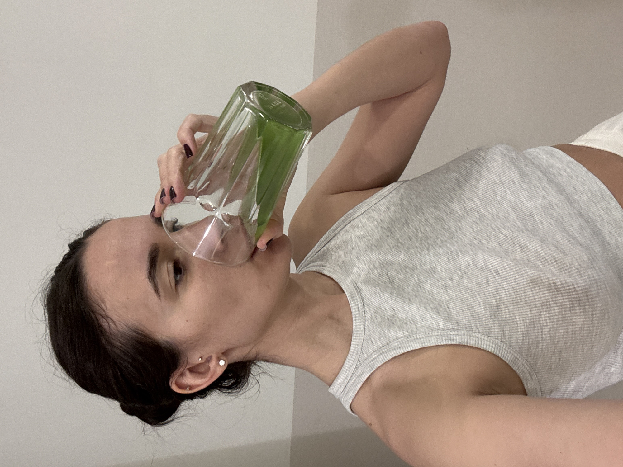 A woman with dark hair pulled back is drinking from a tall, clear, faceted glass filled with a green liquid. She is wearing a grey ribbed tank top. The background is a plain, light-colored wall. There is no indication of the location being Chaikovsky, Russia.