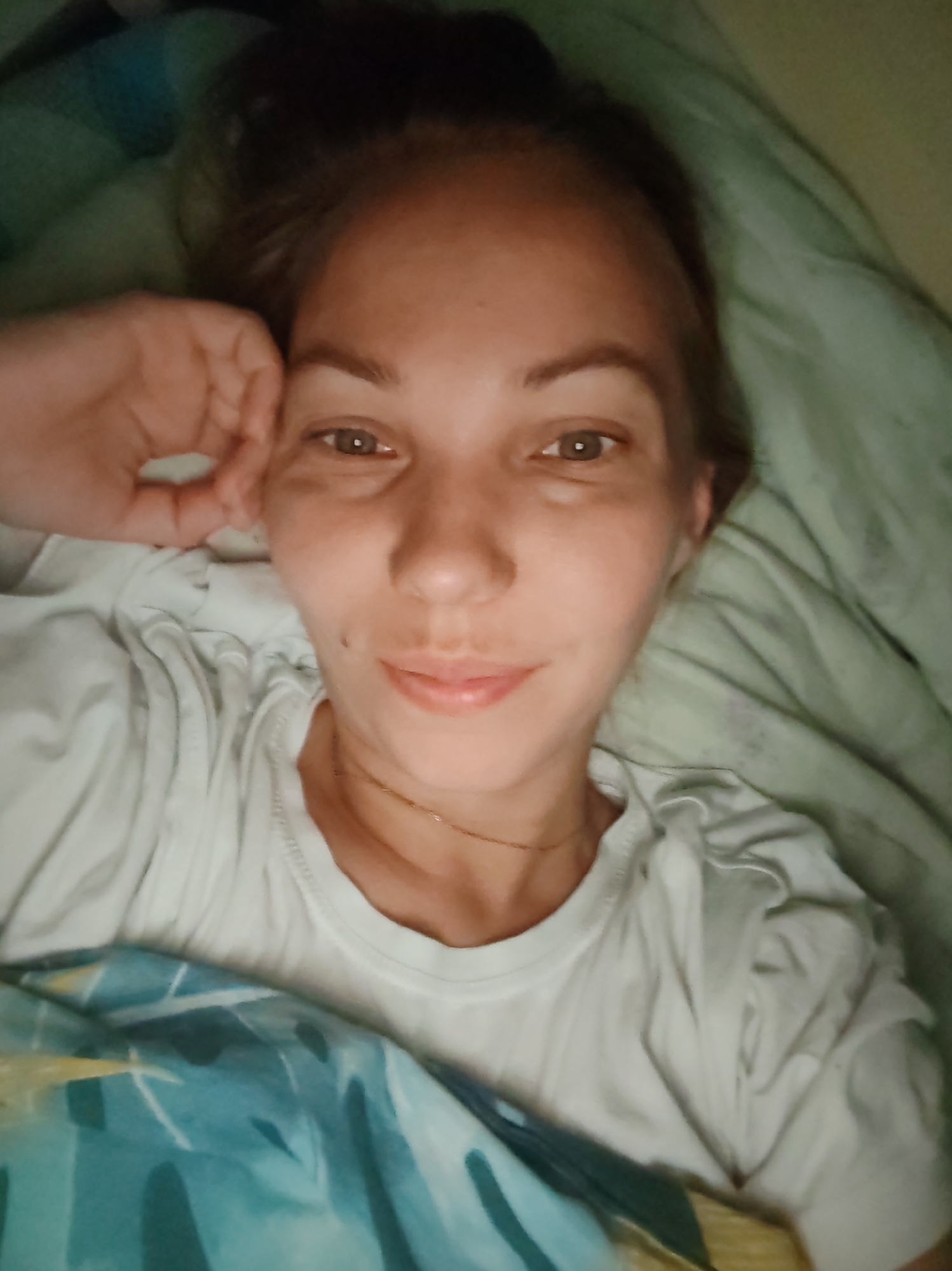A woman with fair skin and light brown hair, pulled back into a bun, lies in bed with a hand resting on her forehead. She is wearing a white t-shirt and a thin gold chain necklace. The background consists of rumpled green and blue bedding. The scene appears to be indoors, likely a bedroom. The lighting is dim, suggesting it may be nighttime or early morning. The image does not contain information to determine the city of Чухлэм, Russia.