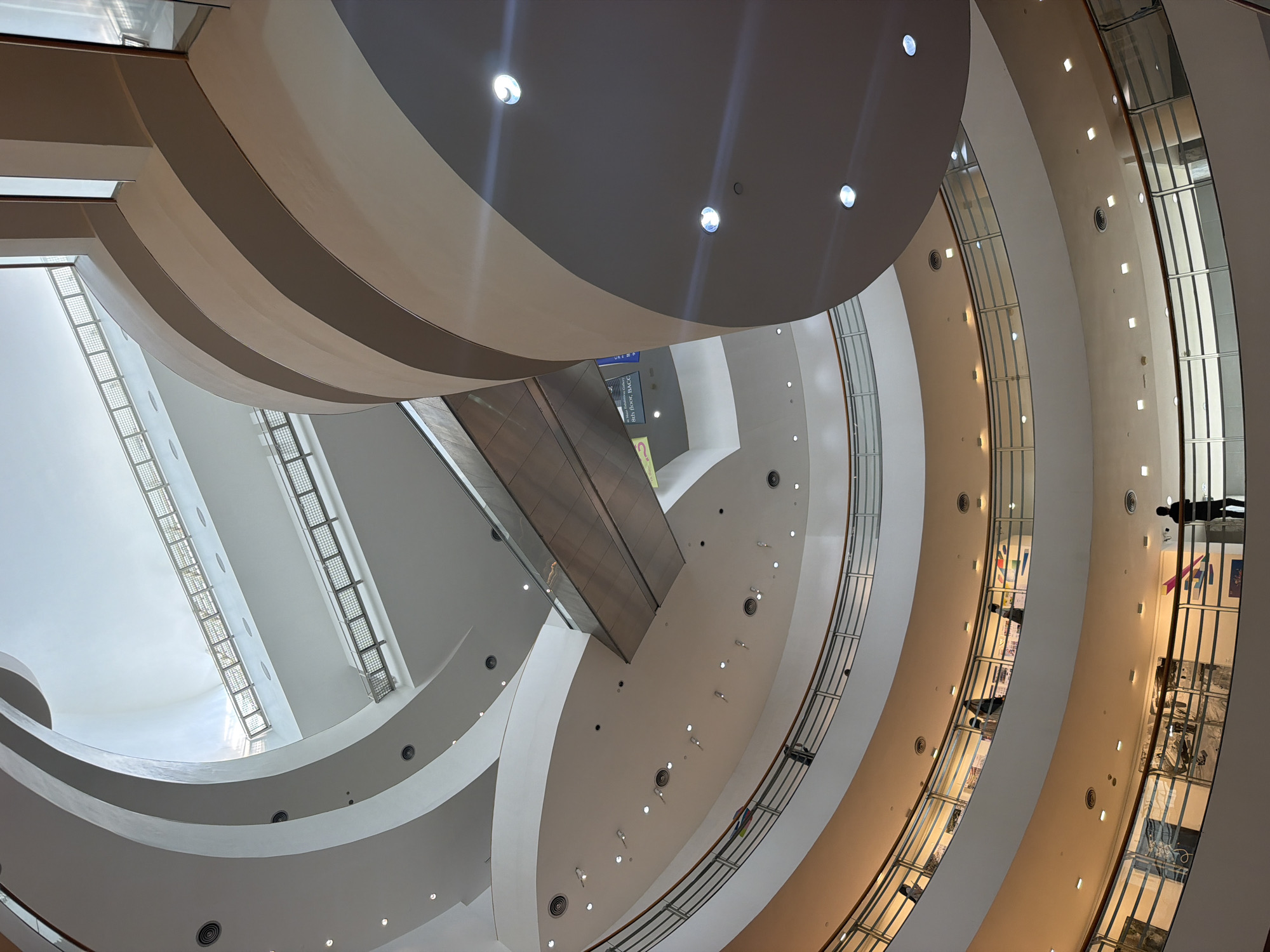 The image depicts the interior of a multi-level shopping mall in Bangkok, Thailand. The architecture features a central atrium with spiraling ramps and escalators. The lighting is a mix of natural light from a large skylight at the top and artificial lighting from numerous circular and square fixtures embedded in the ceilings and walls. Several levels of walkways and balconies with glass railings are visible, with a few individuals browsing or standing. In the foreground, a person stands on a lower level balcony. The overall impression is one of modern, open-plan design with a focus on natural light and flowing architectural lines.
