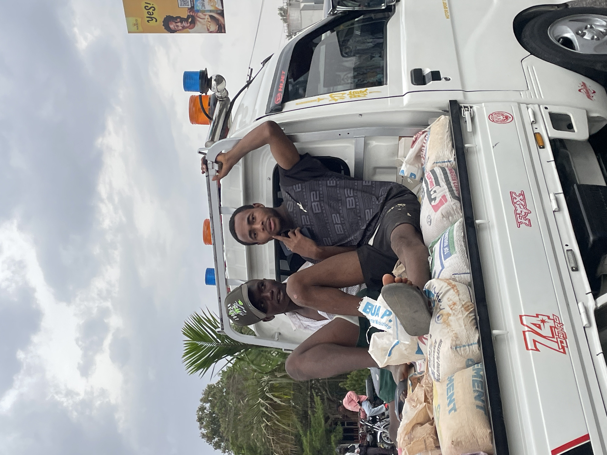 The image depicts two young men riding in the open bed of a white pickup truck, which is visibly laden with numerous sacks of cement. The scene appears to be set on a street or roadside in Jos, Nigeria, under an overcast sky.
The man in the foreground, wearing a dark patterned t-shirt and dark shorts, looks directly at the camera, making a peace sign with his left hand. His bare feet rest casually among the cement sacks. Behind him, another man in a light shirt, dark cap, and dark shorts is also visible, looking towards the camera.
The white pickup truck features custom elements, including blue and orange beacon lights mounted on a rack above the cabin. The truck's bed is filled with sacks, some labeled "CEMENT", "BUA P.", "PLASTER", "FULDING PLA", and "clal Moulding," suggesting they contain building materials.
The background shows a cloudy sky with thick, grey clouds, indicating an overcast day. Palm trees and other green foliage are visible to the left, while a prominent yellow billboard with text and images stands in the upper right. Other blurred vehicles and people can be seen further back, hinting at a busy street environment.
Visible text on the truck includes "HIJET" on a window visor, "MOTOR SPORT" above a window, and a large red graphic "74 TREK" on the tailgate. The pattern on the main subject's shirt includes "B2" and "RARARARI FERRARI FERR". The billboard displays "NASCO", "yes!", and "COINFLA", along with an image of a man and a woman.
The overall activity suggests casual transport of goods, with the men appearing relaxed, as indicated by the peace sign gesture.
