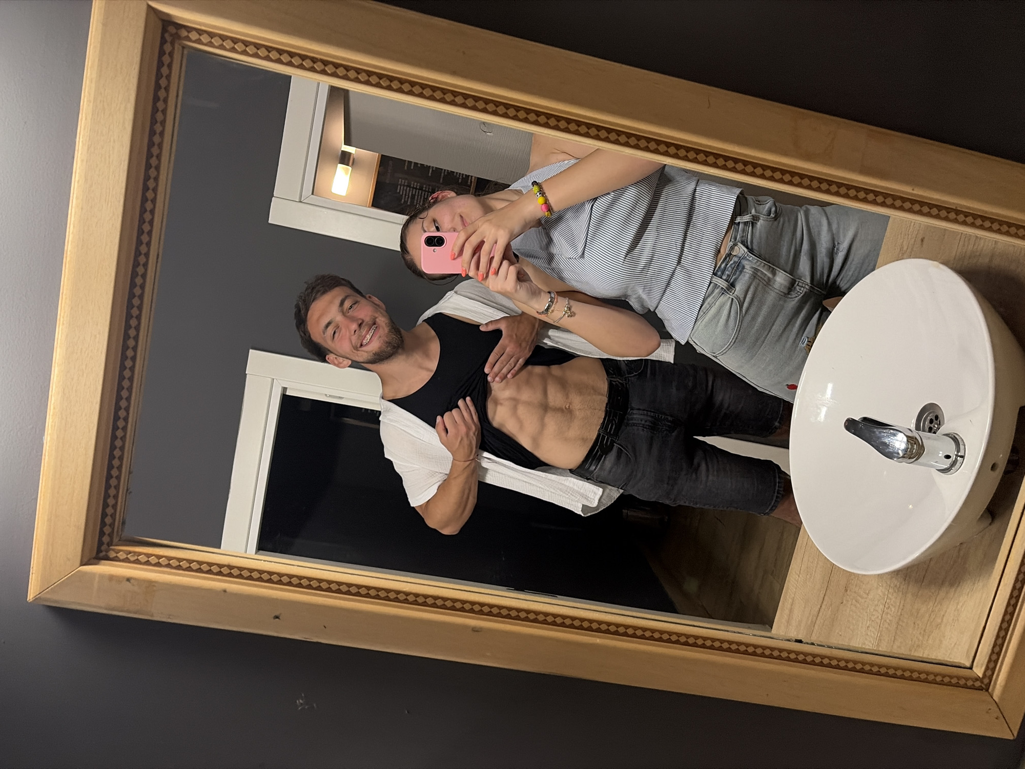A full-length mirror, set within a light-colored wooden frame featuring a repeating dark rhombus pattern, reflects an interior scene in Tsarevo, Bulgaria. The mirror is positioned above a light wood-colored vanity counter, on which a white, round vessel sink with a chrome faucet is centrally placed.
Reflected in the mirror are two individuals: a male on the left and a female on the right. The male has short, dark hair, a beard, and visible braces on his upper teeth; he is smiling directly at the camera. He wears a black tank top under a white, loosely woven short-sleeved shirt, and dark grey denim shorts. His left hand is holding his tank top upwards, revealing a toned abdomen with a visible six-pack. A red string bracelet is on his right wrist.
The female, positioned to the right, is holding a pink-cased smartphone in both hands to capture the image. Her face is partially obscured by the phone, but she appears to be smiling. She has dark hair, likely pulled back, and her fingernails are painted orange. She wears a light blue and white striped halter-neck top and light blue distressed denim jeans. Her left arm displays multiple bracelets, including a silver-toned chain bracelet with a charm and a colorful beaded bracelet.
The reflected background consists of dark grey walls, with two white doorframes visible behind the subjects. A light fixture is reflected above the doorframe on the right. A section of light-colored wooden flooring is also visible in the reflection below the male's legs.