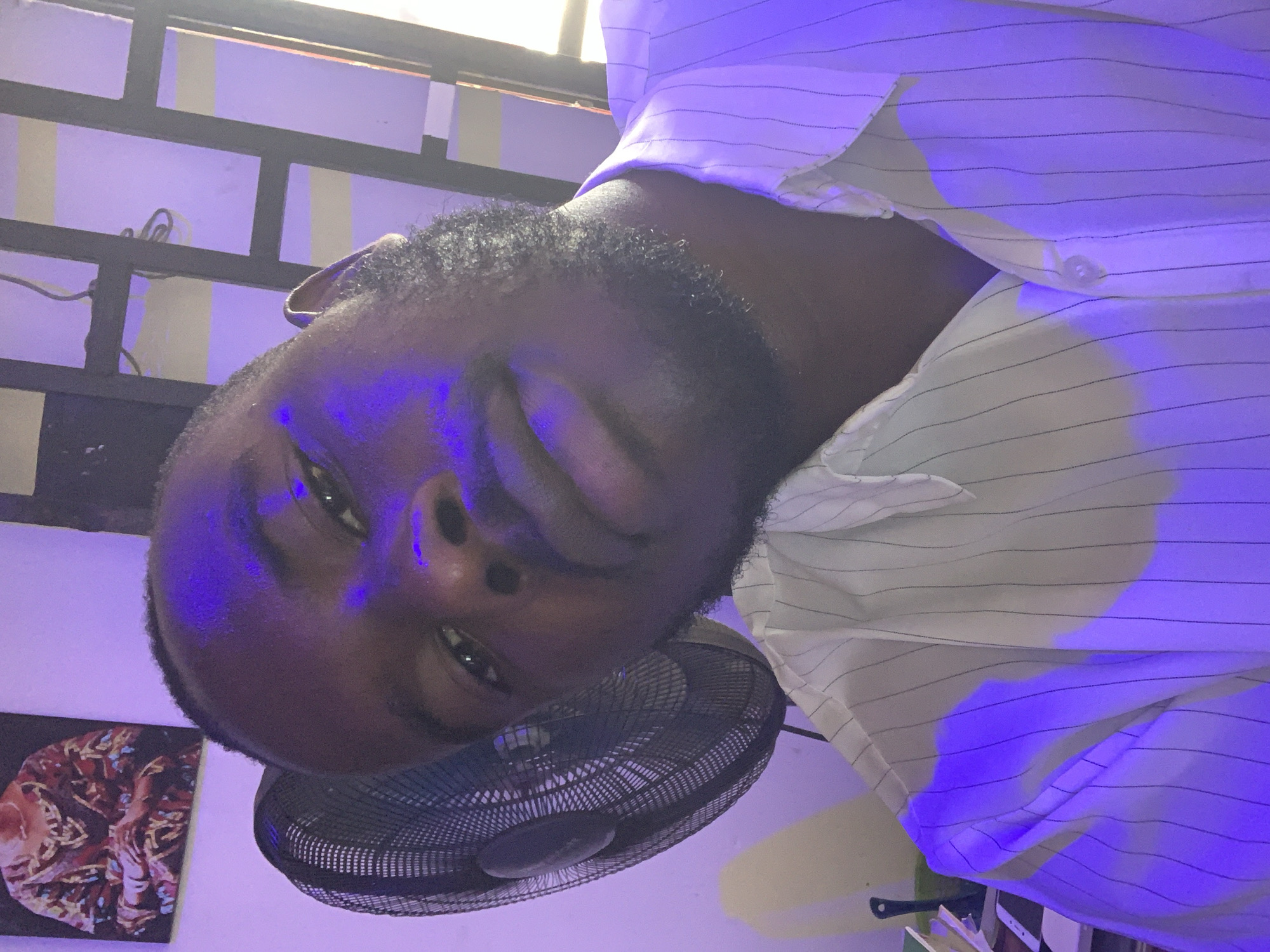An indoor portrait captures a dark-skinned man, possibly in Jos, Nigeria, looking directly at the camera. The image is rotated 90 degrees counter-clockwise. He has a beard and short, dark, curly hair. He wears a white collared shirt with thin dark vertical stripes. A vibrant purple light illuminates his face and the surrounding interior, casting a distinct hue over the scene. One of his hands is partially raised, covering a portion of his face.
Below him, a black electric fan with a mesh guard is visible. The background features a light-colored wall, intensely colored purple by the artificial lighting. A black metal security grating or window frame is seen in the upper part of the image, with wires hanging near it. In the lower left corner, a framed artwork or picture with warm, abstract patterns (red, orange, brown) is mounted on the wall. Another white rectangular object, possibly a phone or book, is partially visible in the bottom right background. The man's expression appears neutral or contemplative. No discernible text is visible in the image. The strong artificial lighting makes the time of day unclear.