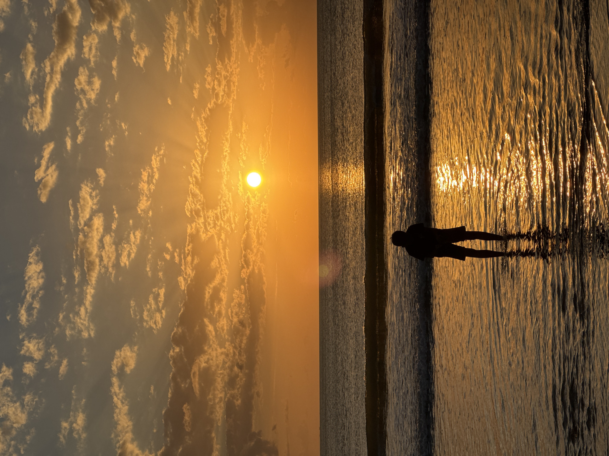 A solitary figure stands in shallow ocean water, facing the setting sun. The sun, a bright orb, is positioned in the upper center of the frame, casting a warm, golden glow across the sky and sea. Rays of light pierce through scattered, orange-tinged clouds. The ocean has gentle waves lapping at the shore, and the water reflects the vibrant colors of the sunset. The figure, silhouetted against the bright background, appears to be wearing shorts and is looking out towards the horizon. The scene is likely located on a beach in Kuta Selatan, Indonesia, a popular tourist destination known for its beautiful sunsets.