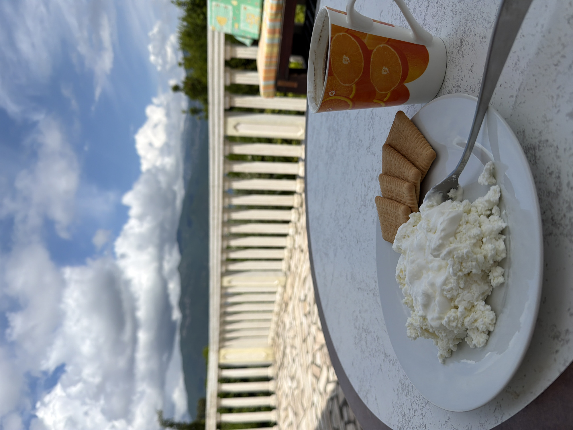 On a table overlooking a partially cloudy sky with mountains in the distance, sits a white plate containing cottage cheese and four rectangular cookies. A silver spoon rests partially in the cottage cheese. A mug with orange slices printed on it stands next to the plate. The location is Krašići, Montenegro. The table is situated on a balcony with white railings.
