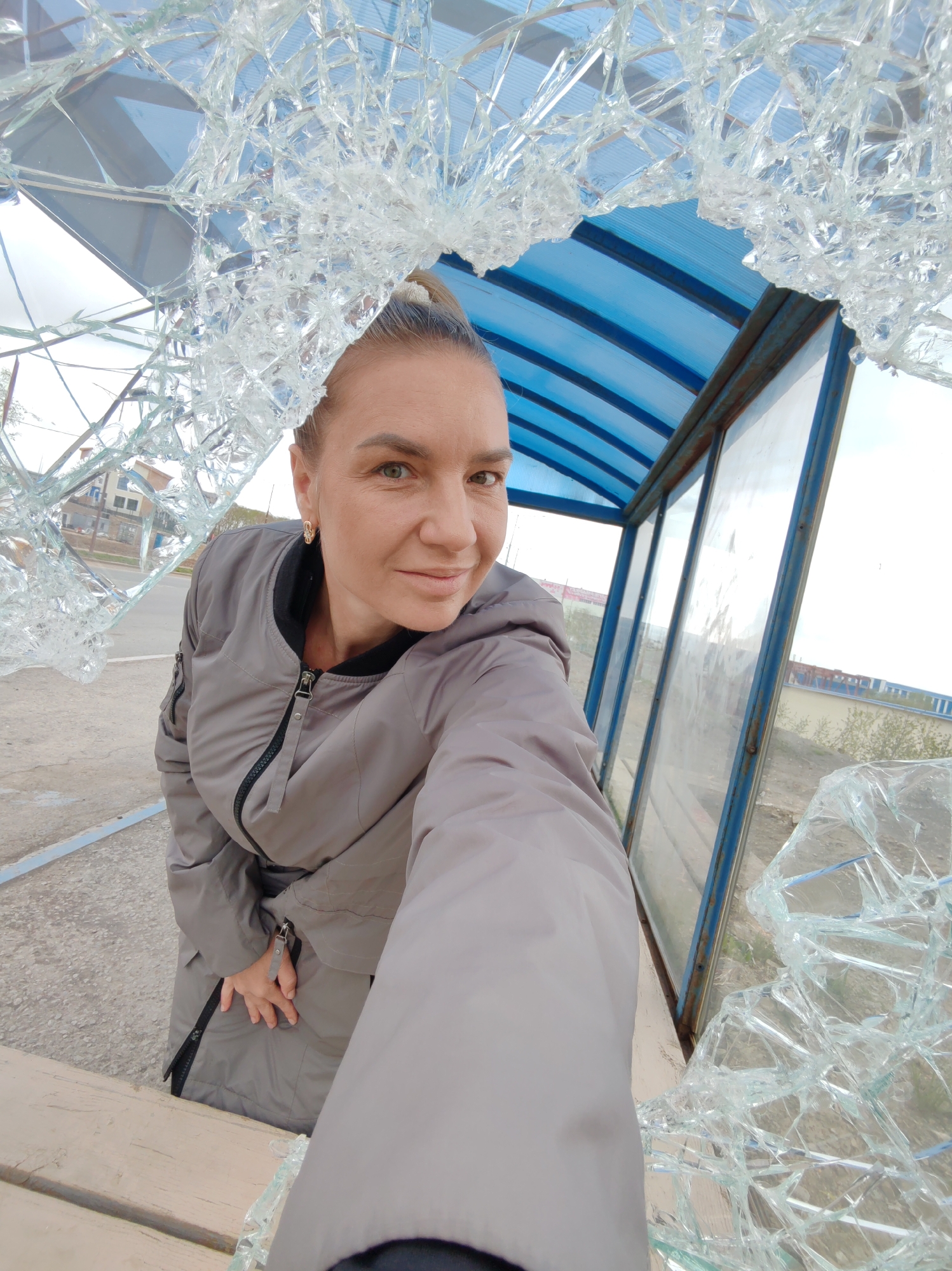 A woman is captured in a selfie, positioned in front of a broken glass structure, possibly a bus stop or shelter, in Petropavlovsk-Kamchatsky, Russia. The woman is wearing a grey jacket and has her hair tied back. She is looking directly at the camera with a slight smile. The foreground of the image is dominated by shattered glass, creating a dramatic frame around the woman. Behind her, the blue roof and window panels of the structure are visible, along with an outdoor environment that appears to be a street or public area with buildings in the background. The overall scene suggests a damaged public amenity and a casual moment captured by the individual.