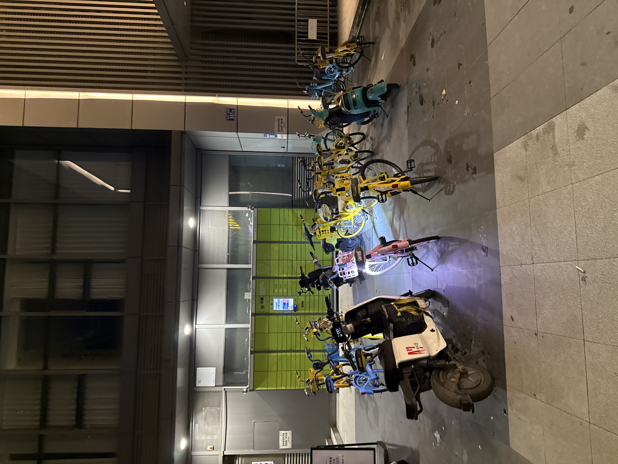 The image shows a street scene in Tianhe District, China, at night. A row of shared electric scooters and bicycles are parked on a sidewalk. Some of the vehicles are yellow, while others are blue and green. A white and black electric scooter with a "H7" logo is in the foreground. The buildings in the background appear to be modern with metal grates and glass windows. The lighting suggests it is nighttime, and the weather appears to be clear. There are no people visible in the image. The scene depicts a common urban amenity in China. Text visible on a blue sign reads "081". Another sign on a green structure is partially visible with Chinese characters.