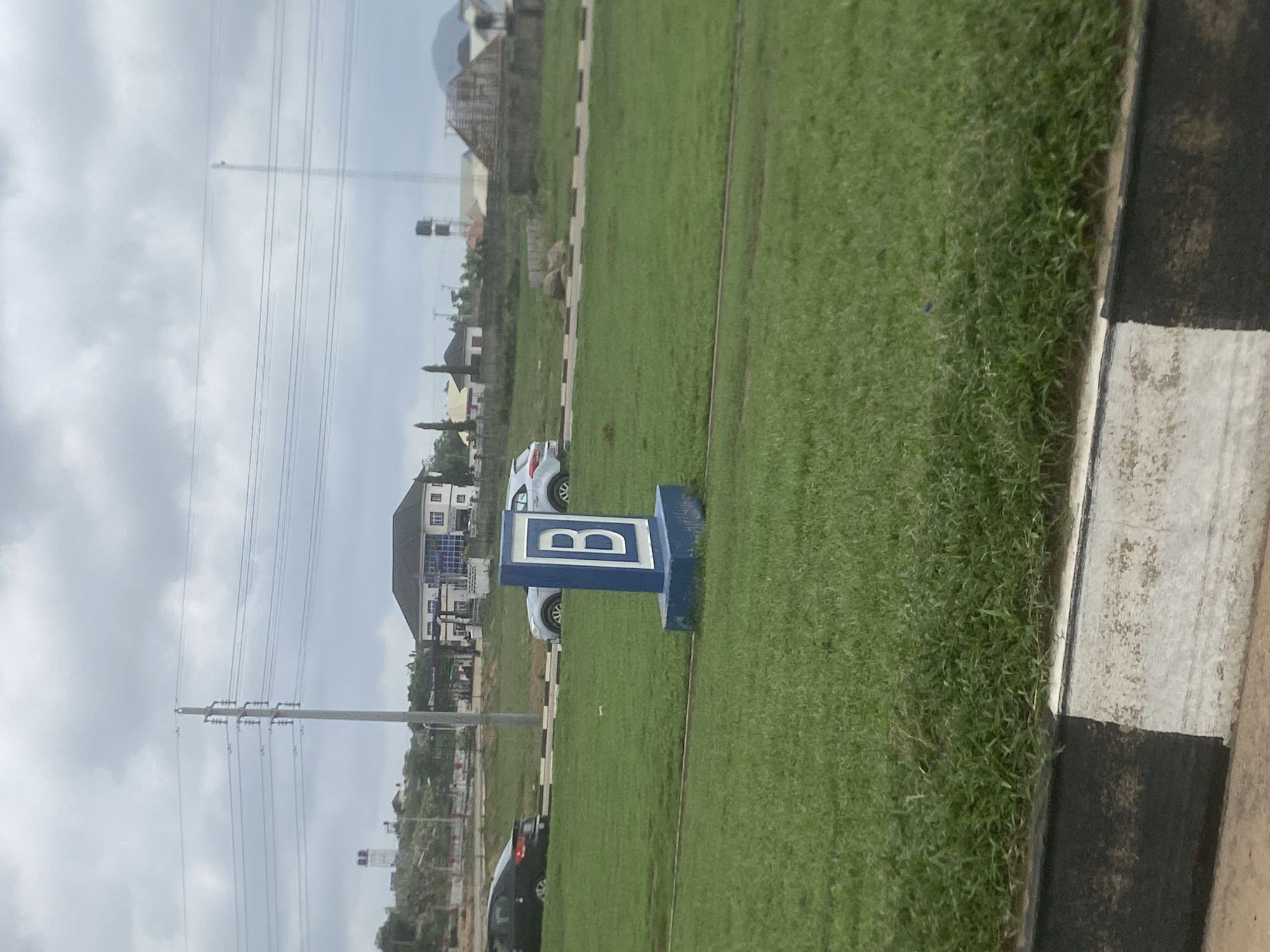 The image presents a wide green field under an overcast sky during daytime in Gosa, Nigeria. In the midground, a prominent blue and white rectangular sign stands on the grass, clearly displaying the capital letter "B". To the right, a curb with distinct black and white stripes borders the grassy expanse.
In the background, several residential buildings and houses are visible, featuring various architectural styles, some with multiple stories. A few cars, including white and dark models, are parked near these structures. Further into the distance, a tall utility pole with numerous power lines stretches across the sky, and another water storage tower can be seen. The scene suggests a developed or developing area with a mix of open green spaces and built infrastructure. No human activity is visible.