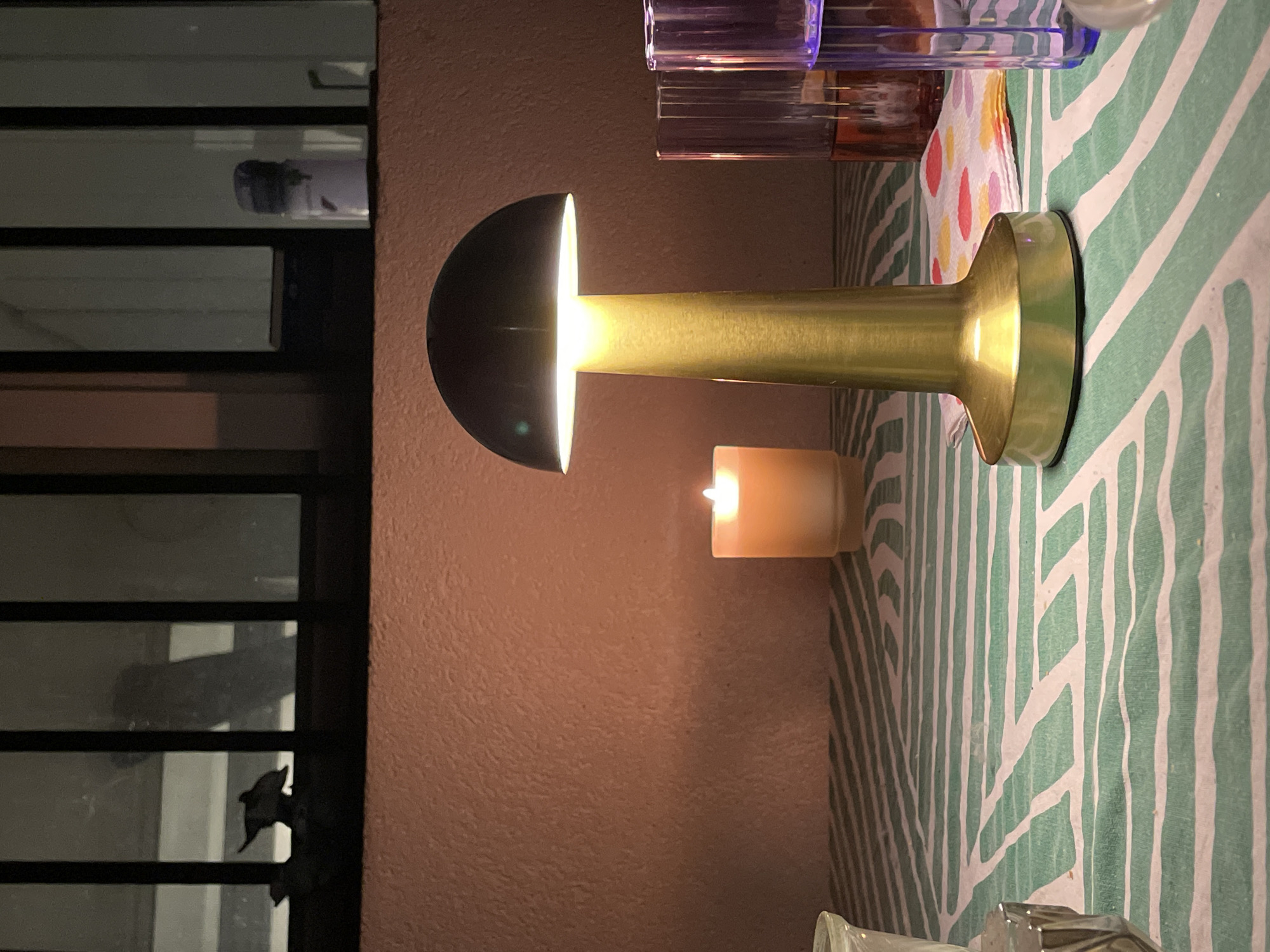 The image depicts an indoor scene, potentially a room in a building, as indicated by the presence of a window with a metal frame and panes on the left side. Outside the window, vague shapes and shadows suggest a dimly lit environment, possibly urban. Inside, the scene is illuminated by a lamp with a half-spherical shade, casting a bright light onto its brass-colored body. A small, lit candle sits nearby, contributing to the warm lighting. The table beneath the lamp and candle is covered with a tablecloth featuring a green and white zigzag pattern. Other objects, including drinking glasses, are present on the table. It is not possible to determine the location in the city of Sofia, Bulgaria. There are no people present in the image.