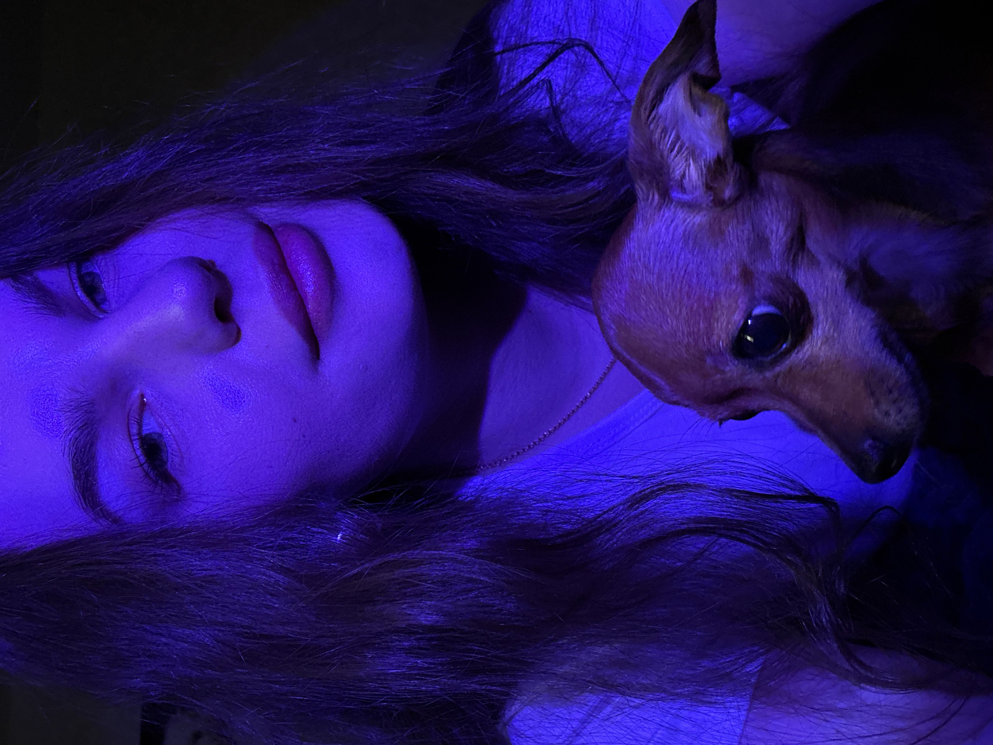 A medium shot features a woman and a brown, smooth-coated dog under blue lighting. The woman lies on her back, with her head tilted to the right. Her long, dark hair is spread around her head and shoulders. Her eyes are open, and she is looking slightly to the left. Her lips are full and appear to be tinted with a reddish-pink hue. The dog is positioned to the right of the woman's head, with its head close to hers. The dog is facing forward. The background is dark and indistinct. The location is not discernable from the image.