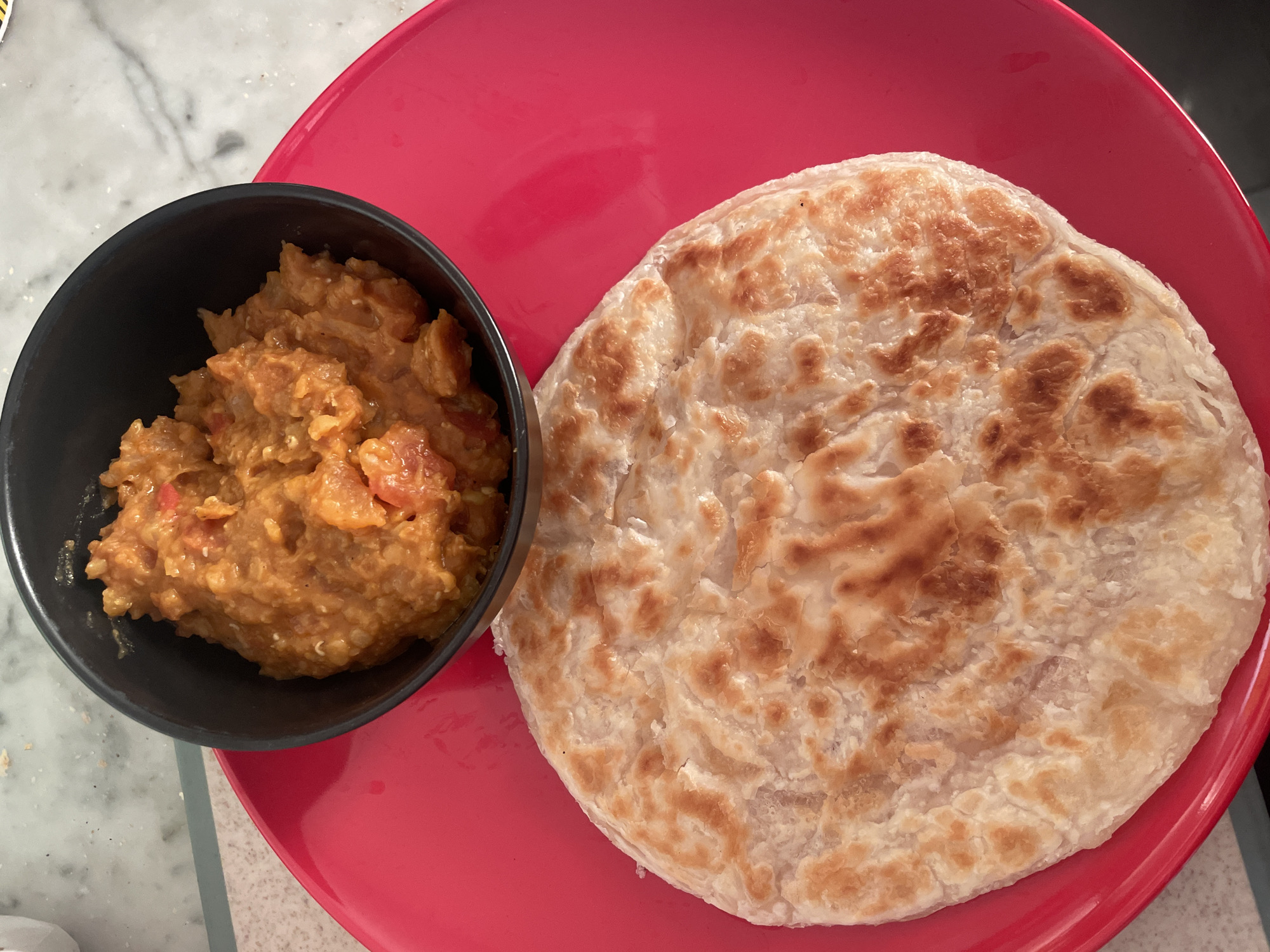 A top-down, medium shot captures a plate of food on a marble-patterned countertop. The plate is bright red and round, with a slightly raised rim. Dominating the foreground is a large, circular, flaky flatbread, lightly browned in places. In the upper right quadrant of the plate sits a black bowl, filled with a thick, orange-brown stew or curry. The stew appears to be made of small grains and chunks of red vegetables, possibly tomatoes. The background shows a marble countertop with subtle gray veining. The scene is static and appears to be indoors, likely in a kitchen or dining area. No people are visible. The image does not contain any information that would allow for the identification of a specific location in Melbourne, Australia.