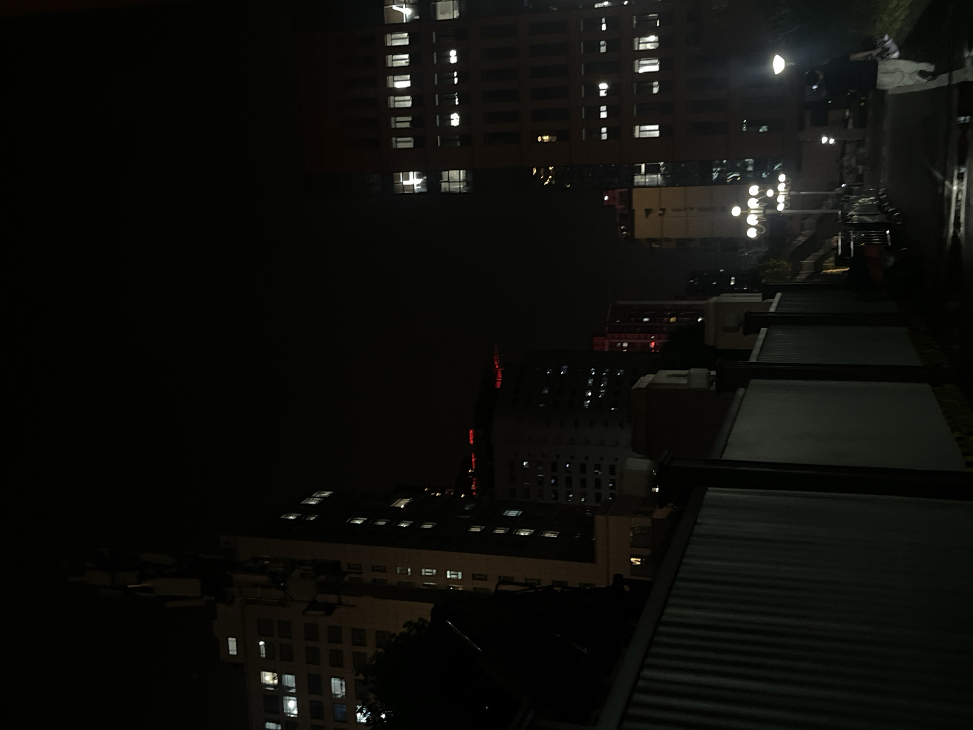 The image depicts a nighttime urban scene in Yuzhong District, China. Tall buildings with illuminated windows dominate the background. In the foreground, a dark, corrugated metal fence lines a pathway. Several streetlights illuminate the path, casting a glow on the wet asphalt surface. A lone figure, dressed in light-colored pants and a dark shirt, walks away from the camera down the path, carrying what appears to be a bag. The overall atmosphere is dark and somewhat subdued, with the artificial lights of the city providing the primary illumination.