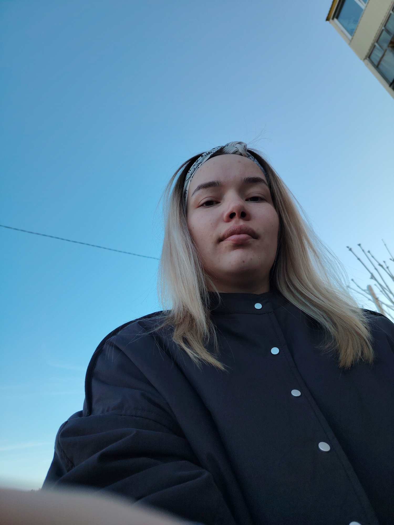 A young woman stands outside in what appears to be the Markovskii district of Russia, as a building is visible in the background. The sky is a clear, light blue. She is wearing a dark, button-down jacket and a patterned headband. Her blonde hair is loose around her shoulders. The photograph is taken from a low angle, looking up at her.