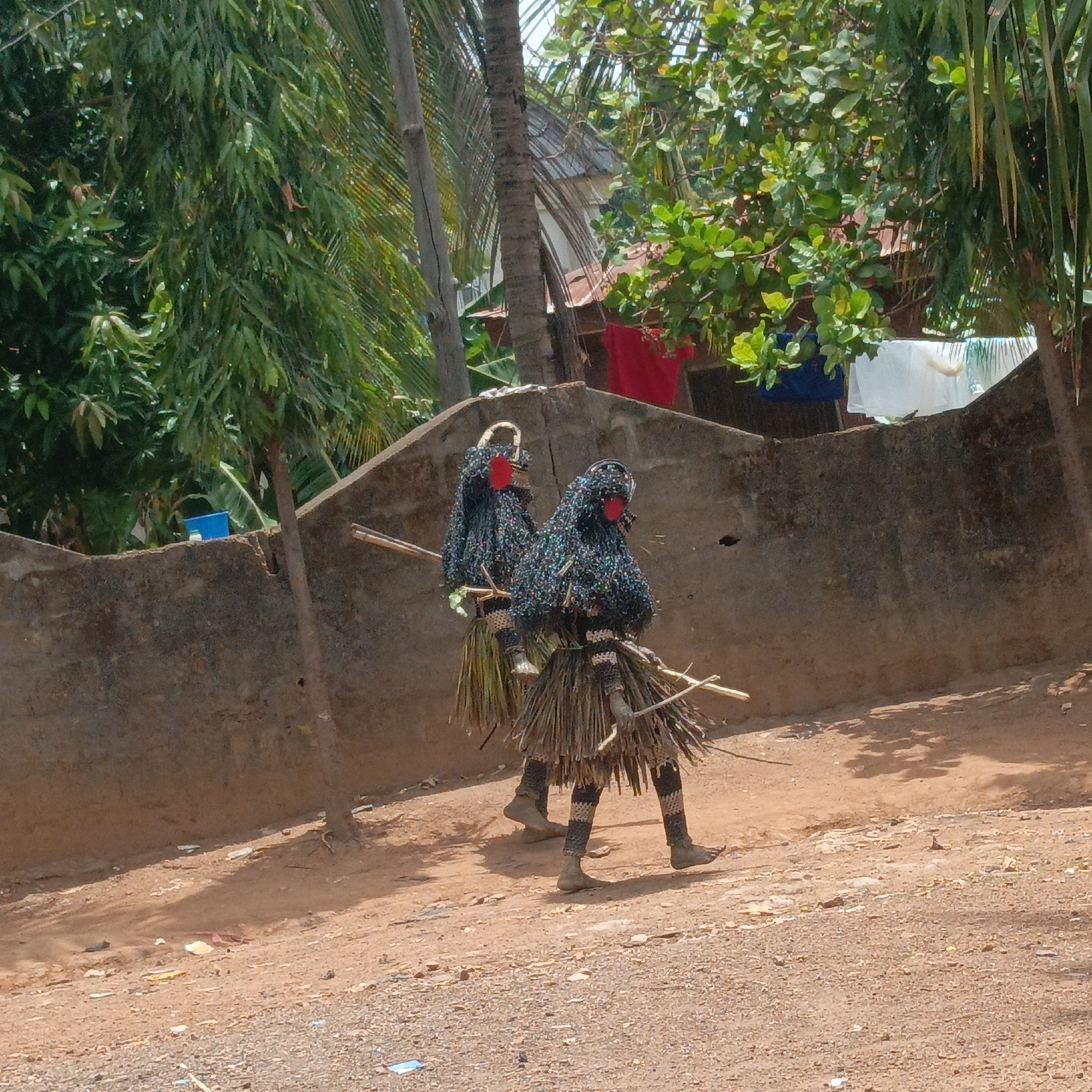 Two people dressed in traditional masquerade costumes are walking on a dirt path in Akwanga, Nigeria. The costumes consist of black, patterned outfits with red facial coverings and straw skirts. They are each carrying sticks. A low concrete wall is behind them, with laundry hanging on the other side. Trees surround the area.
