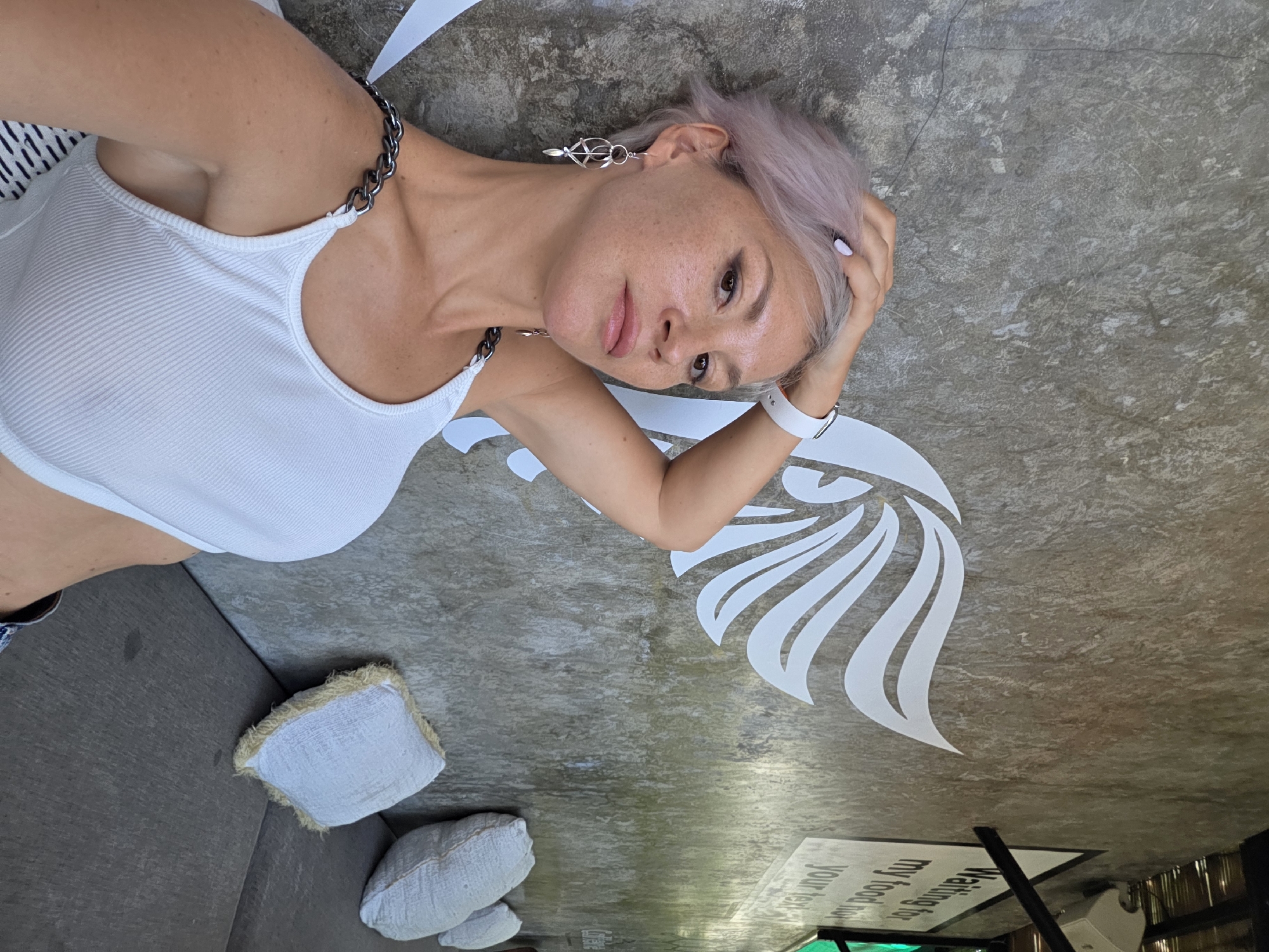 The image is a vertically-oriented, high-angle shot of a woman lying on a concrete surface. The woman has short, light purple hair, fair skin with freckles, and wears a white tank top with a chain strap. She also has a white wristwatch on her left wrist and dangling silver earrings. Her left hand is resting on her head.
The concrete surface has a light gray hue with mottled textures. In the upper-right portion of the frame, there is a large white decal depicting a stylized wing or feather design. The lower-right corner shows a sign with green text, which includes the phrase "Waiting for my food." In the lower-left part of the frame, there are three small white cushions with fringed edges.
The location is in North Kuta, Indonesia.