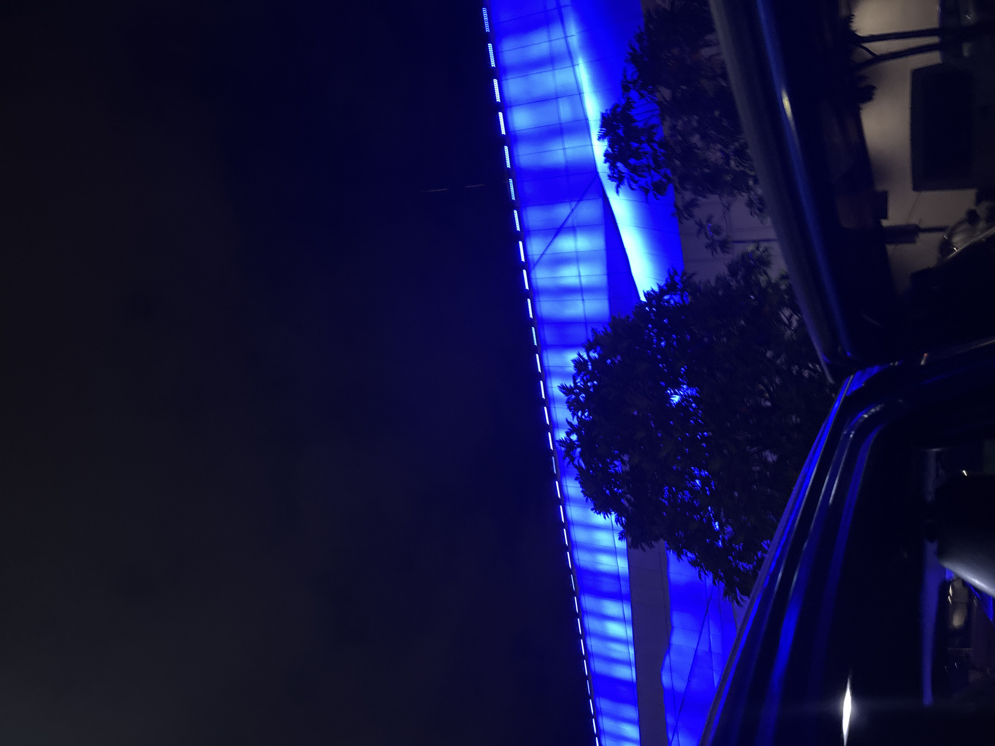 The image captures a night scene in Davao City, Philippines. A large, modern building is illuminated with vibrant blue lights, creating a striking contrast against the dark sky. Trees, silhouetted by the blue glow, are visible in front of the building. In the foreground, the dark silhouette of a vehicle's interior, possibly a car or bus, is visible, with some reflections of light on its surfaces. The overall impression is one of urban nightlife and architectural illumination.