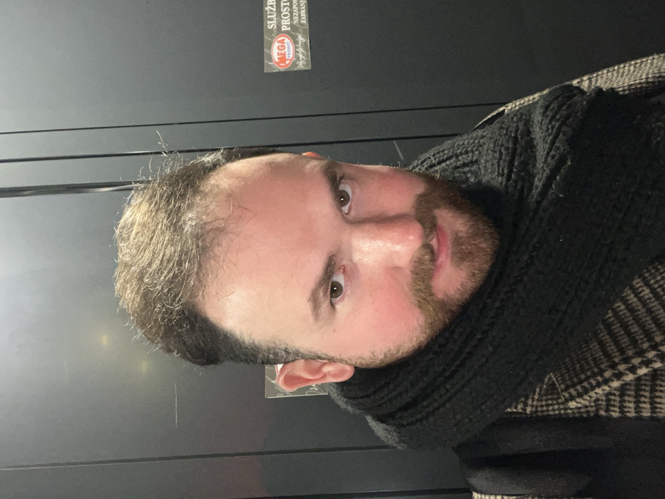 A fair-skinned man with brown hair and a beard faces the camera. He wears a black turtleneck sweater and a checkered coat. The background is a dark grey wall, with a sign visible in the upper left corner. The image is slightly tilted, with the man's head turned at an angle. His expression is neutral, and he appears to be in an indoor setting.