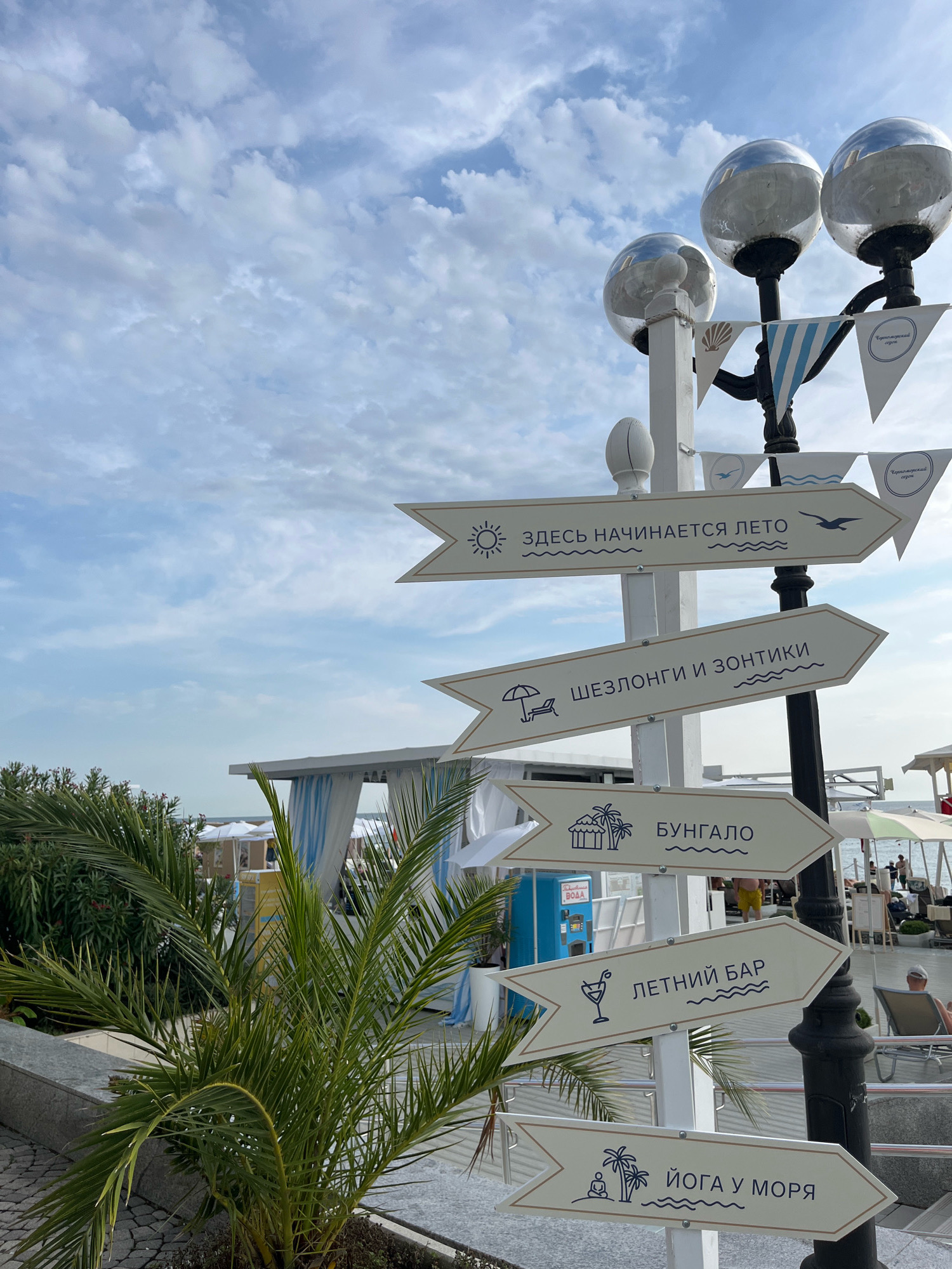 The image depicts a directional signpost on a cloudy day, likely located in a coastal area of Sochi, Russia. The signpost has multiple arrow-shaped signs, each with a pictogram and text in Russian. The signs point to various amenities and locations. The topmost sign reads "ЗДЕСЬ НАЧИНАЕТСЯ ЛЕТО" (Summer Starts Here) with a sun and a bird icon. Below it, a sign indicates "ШЕЗЛОНГИ И ЗОНТИКИ" (Sunbeds and Umbrellas) with an icon of a sun lounger and an umbrella. The next sign points to "БУНГAЛО" (Bungalows) with a depiction of palm trees and a hut. Further down, "ЛЕТНИЙ БАР" (Summer Bar) is indicated by a cocktail glass icon. The bottom sign reads "ЙОГА У МОРЯ" (Yoga by the Sea) with an icon of a person meditating and palm trees.
In the background, there are beach cabanas with white and blue striped curtains, white umbrellas, and lounge chairs, suggesting a resort or beach club setting. Palm trees and other greenery are visible in the foreground and to the left of the signpost. A streetlamp with three spherical globes stands behind the signpost. The sky is filled with scattered clouds. In the distance, the sea and a few people on lounge chairs can be seen. The overall impression is a well-equipped beach or resort area, likely a popular tourist destination.