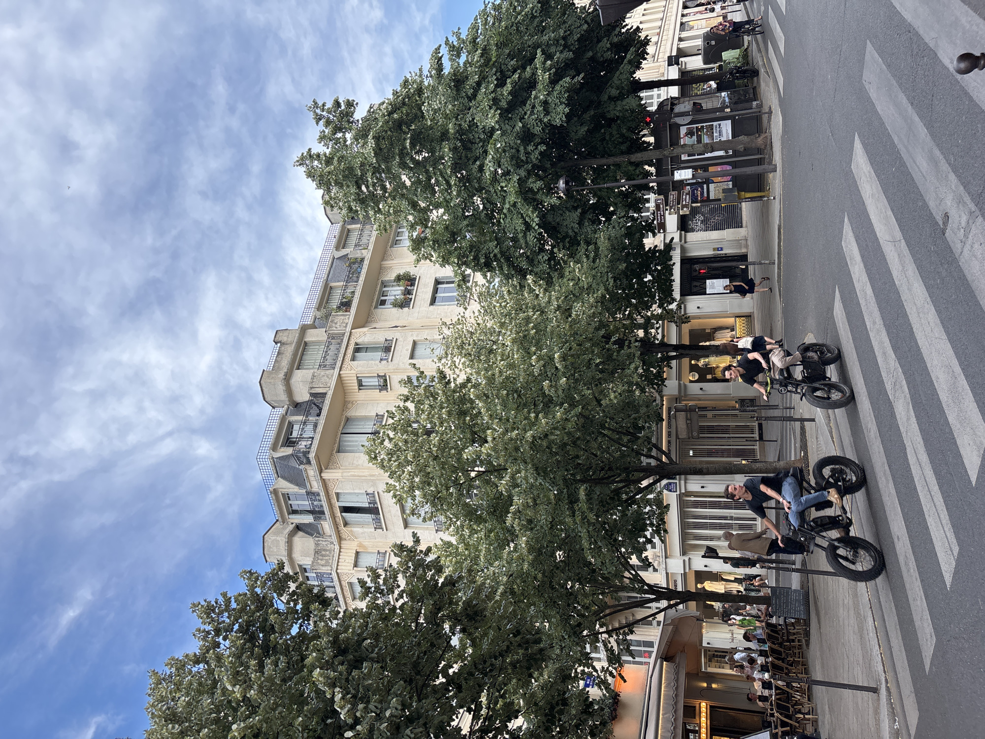 The image depicts a street scene in Paris, France, with a building in the background and two individuals riding electric motorcycles in the foreground. The building appears to be Haussmannian architecture, characterized by its mansard roof and ornate facade, typical of Parisian apartment buildings. Trees line the street, providing shade and a touch of greenery. A crosswalk is visible, indicating a pedestrian crossing. One rider, wearing a black t-shirt and blue jeans, is on the left, while the other, in a black shirt and light-colored pants, is on the right. Both are facing forward, seemingly riding towards the viewer. In the background, patrons are seated at tables outside a cafe, and shops line the street. The sky is partly cloudy, with a mix of blue and white. The overall impression is a lively urban environment with a blend of traditional architecture and modern transportation.