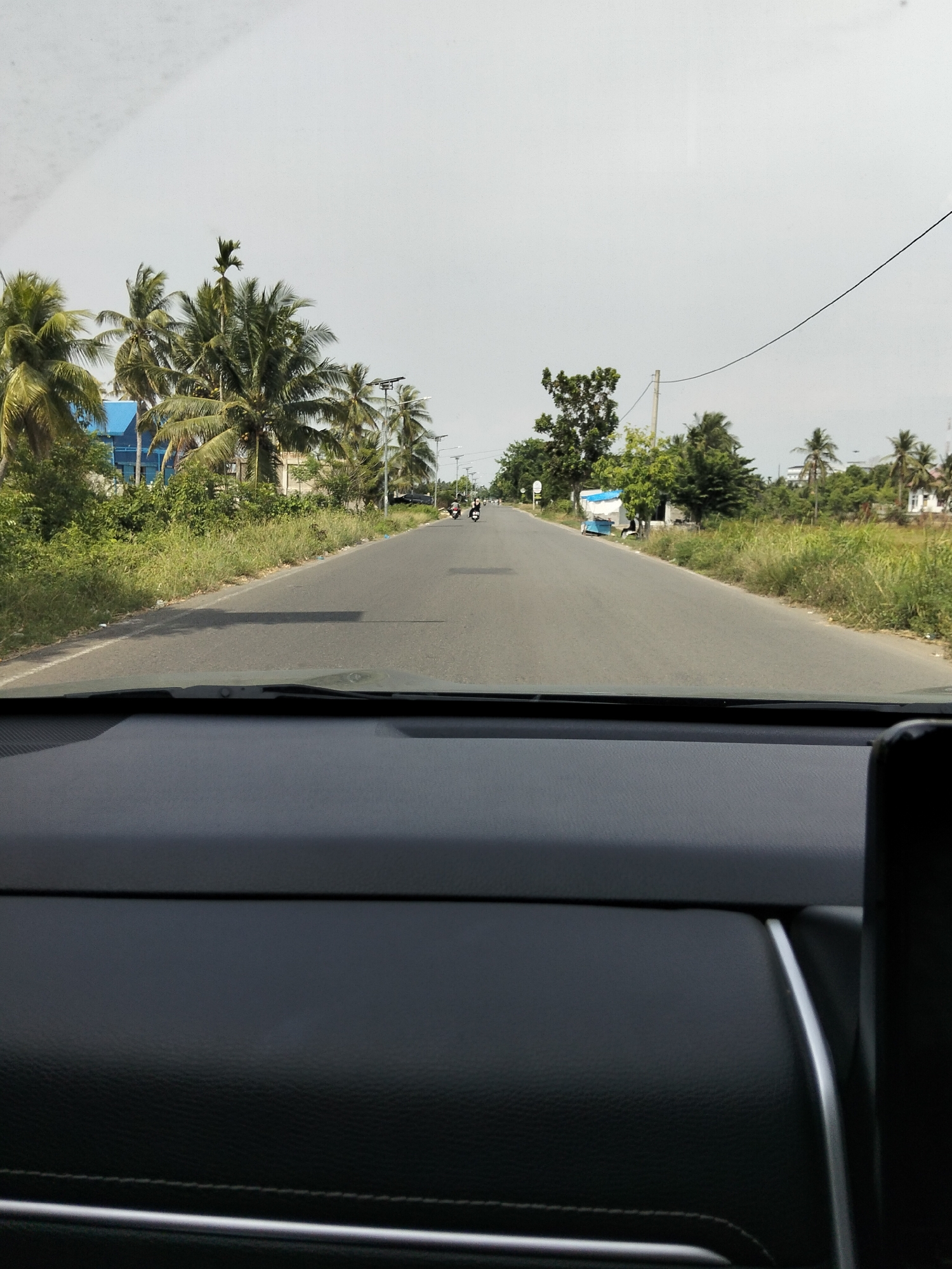 The media file depicts a view from inside a vehicle, looking out through the windshield at a two-lane road. The scene is an outdoor, rural setting in what appears to be a tropical or subtropical region, given the presence of numerous palm trees. The road stretches into the distance, with the right lane appearing to be empty and the left lane showing two motorcycles traveling away from the viewer.
On the left side of the road, there are lush green bushes and trees, with a blue building visible behind them. Further down the road on the left, more trees and vegetation are present. On the right side of the road, there is a stretch of tall, dry grass, and further in the distance, there are more trees, houses, and what looks like a small structure with a blue tarp. Electrical poles and wires are also visible along the roadside. The sky is overcast, suggesting a cloudy day.
The time of day is not explicitly clear, but the ambient light suggests it is daytime. The overall impression is one of traveling through a quiet, possibly somewhat developing, area. There is no visible text within the image. The activity is simple travel along a road in a rural landscape.