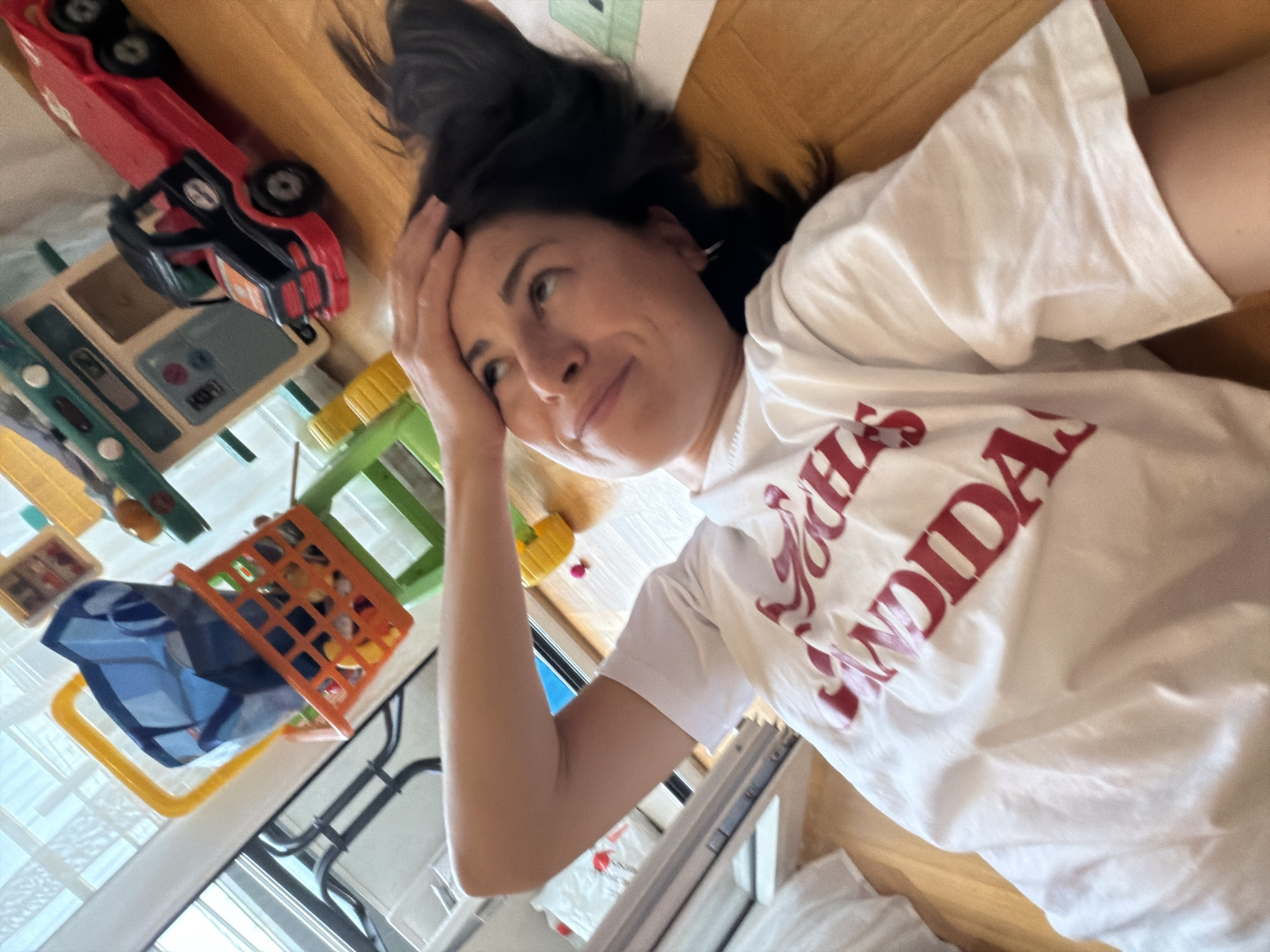 A woman lies on a wooden floor, her head resting on her hand. She looks up and left. Her hair is black. She is wearing a white t-shirt with red text. To her left are toys, including a red toy truck, a wooden kitchen set, a green and yellow toy, and a blue and orange toy basket. A window is also visible to the left. The location cannot be confirmed as Budva, Montenegro from the image alone.