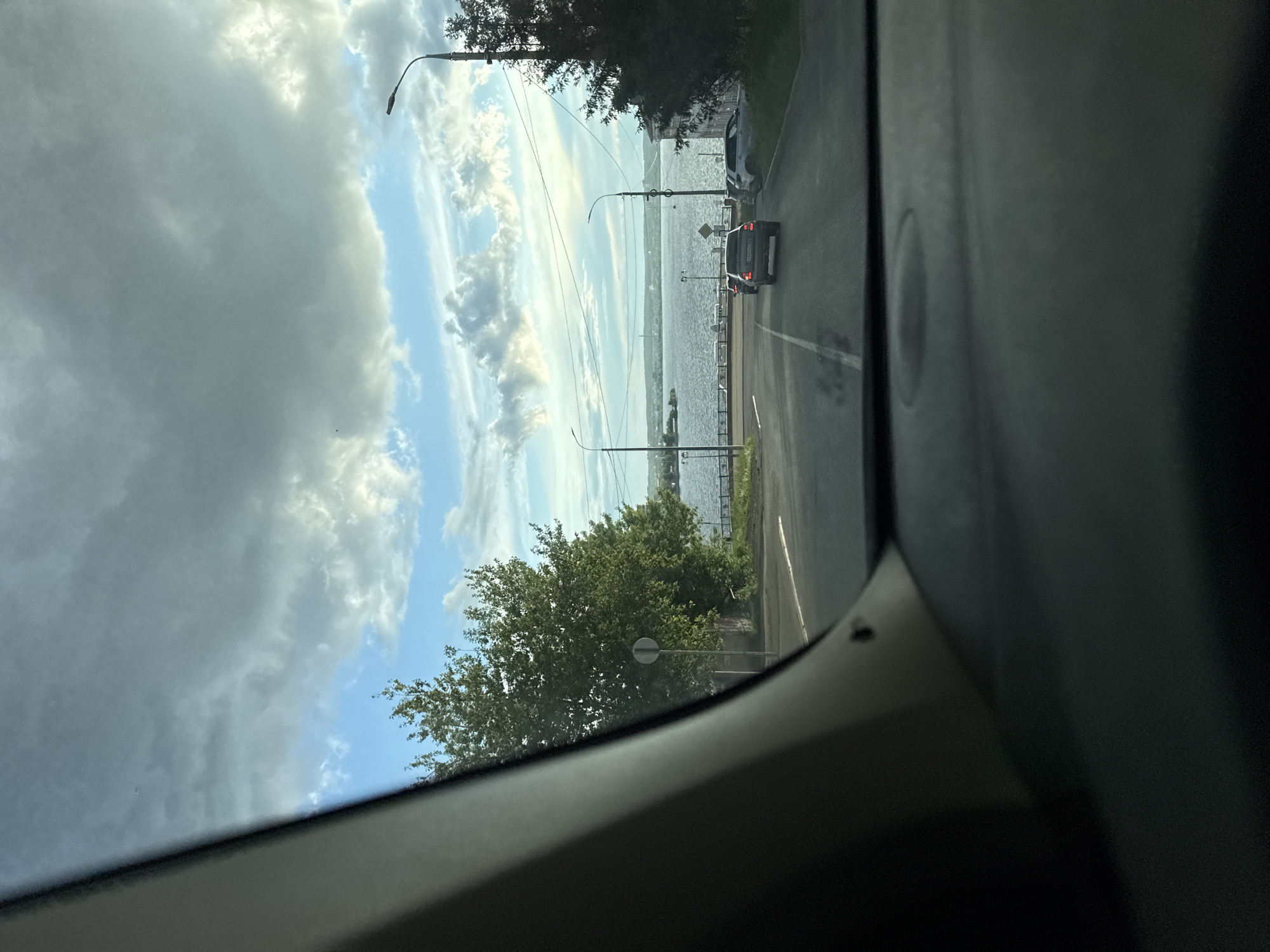 The photograph, taken from inside a vehicle, depicts a road running alongside a body of water, most likely the Izhevsk Pond in Izhevsk, Russia. The road is asphalt and features a car traveling away from the viewer. Lamp posts line the road, interspersed with greenery. The sky is partially overcast with a mix of dark and light clouds. The water appears calm. The interior of the vehicle is visible on the sides of the frame.