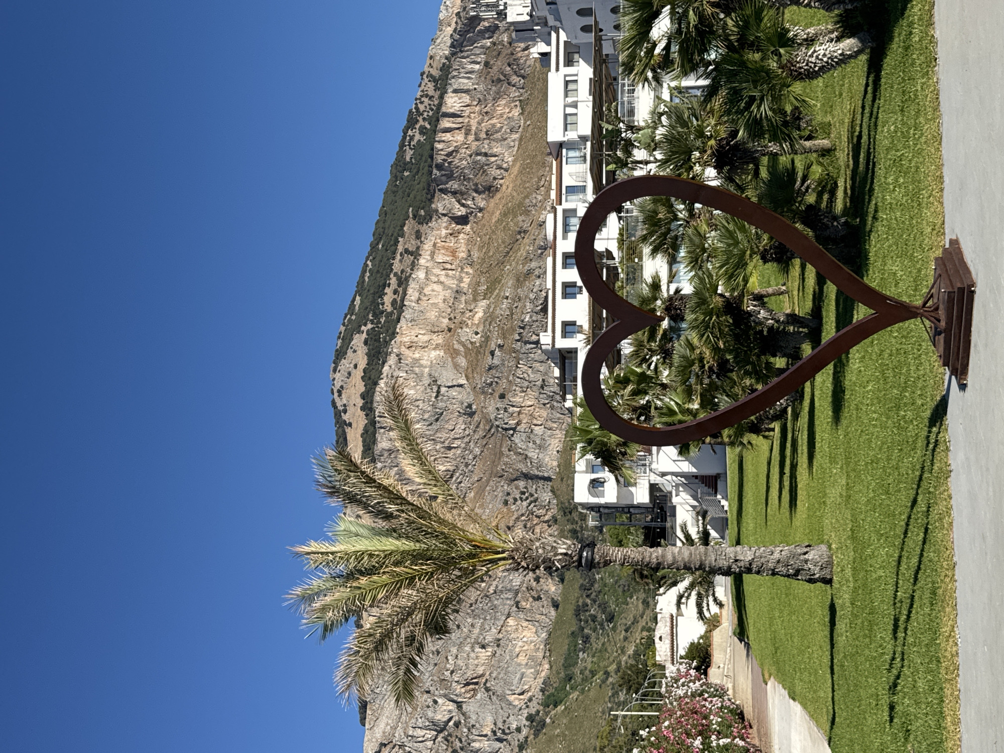 In Isola delle Femmine, Italy, a rust-colored heart sculpture stands on a square base in the foreground of a manicured green lawn. Behind the sculpture, a tall palm tree is visible on the left, its fronds reaching towards a clear blue sky. In the mid-ground, a modern white building with multiple balconies and windows is situated, surrounded by several smaller palm trees. Behind the building, a rugged, rocky mountain with sparse vegetation rises dramatically. To the left of the building, a path lined with pink flowering bushes leads towards the structure. The scene is bathed in bright sunlight, casting distinct shadows on the grass.