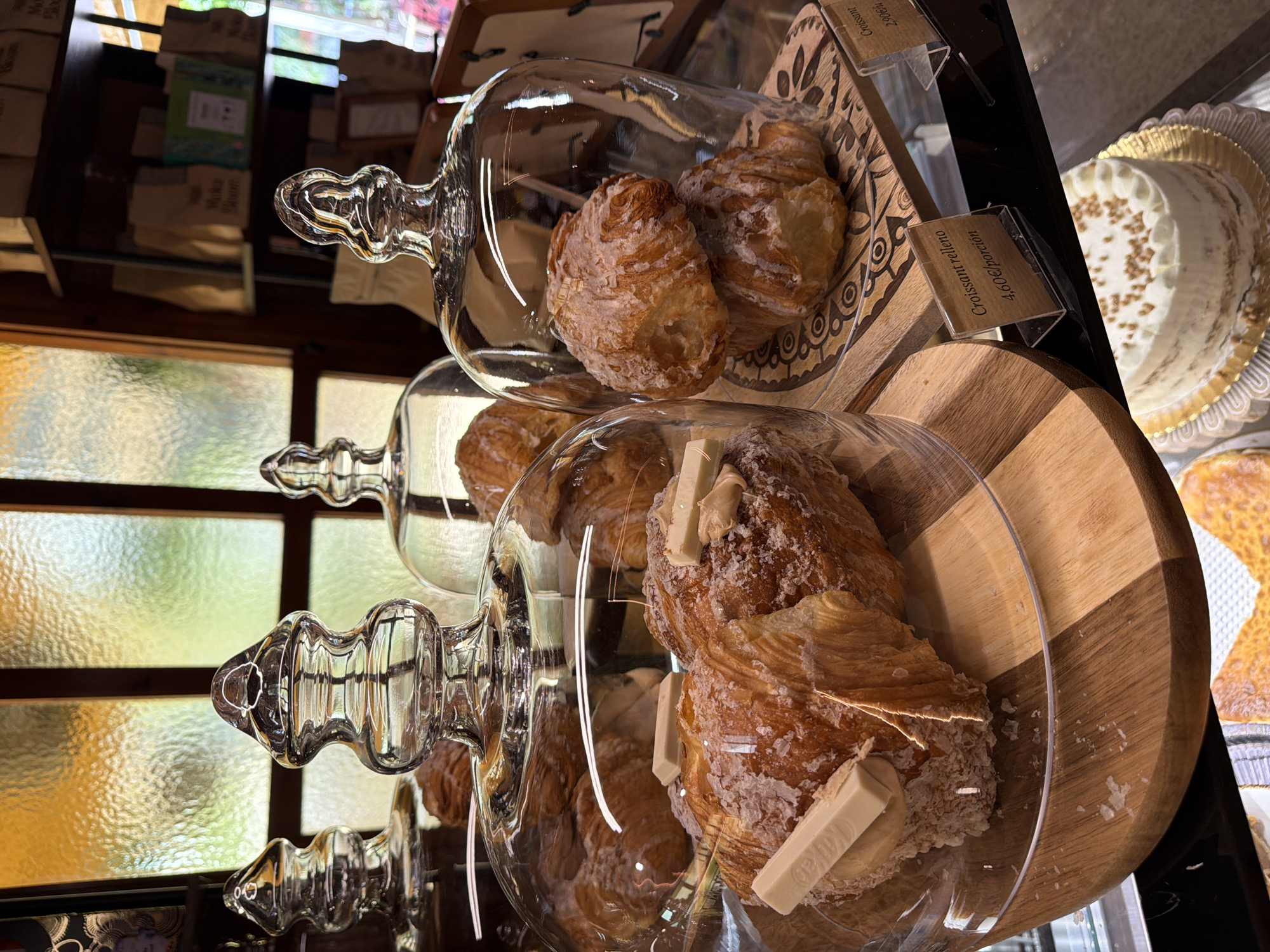 The image depicts a close-up view of a bakery display case, likely in Benidorm, Spain, showcasing several croissants. In the foreground, a large croissant is prominently displayed under a glass dome. It is generously sprinkled with what appears to be sugar and is topped with white chocolate pieces and a dollop of cream or frosting. To the right, another glass dome covers two smaller croissants, also coated in a sugar-like topping. Further back, more croissants are visible under additional glass domes. The croissants are arranged on wooden stands. A price tag for "Croissant relleno" at "4,60€/porcion" is visible in the lower right quadrant of the image. In the background, shelves are stocked with packaged goods, and a large window with textured glass allows diffused light to enter, creating a warm and inviting ambiance. A partially visible cake with white frosting and a sprinkle of toppings is present in the lower portion of the image, suggesting a variety of baked goods are offered. The overall scene is one of abundance and tempting treats within a well-lit establishment.
