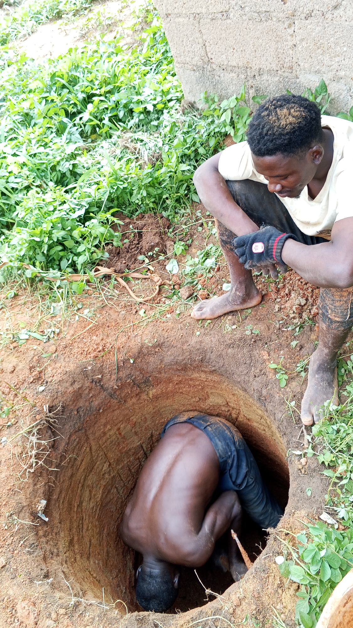 A man is digging a well in Bukuru, Nigeria. The well is in a yard with green vegetation and is dug into red soil. One man is inside the well, bent over and working, while another man sits at the edge of the well, observing. The man inside the well wears jeans. The man sitting outside wears a light-colored shirt and one glove.