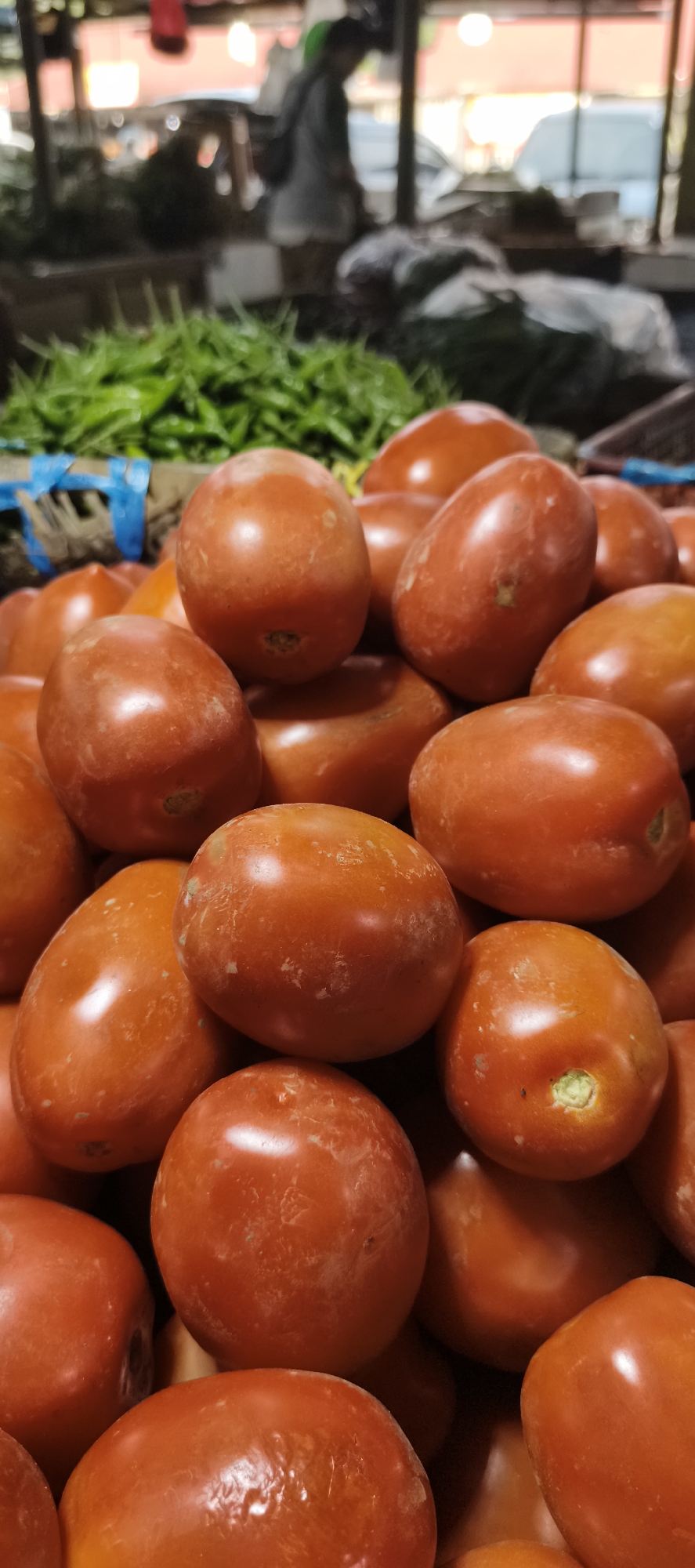 The image shows a close-up of a pile of red tomatoes at a market stall. In the background, blurred figures of people and green vegetables can be seen. The overall impression is of a vibrant, bustling market environment. The location is identified as Jambi, Indonesia, suggesting it is likely an outdoor or covered market setting typical of Southeast Asian cities. The lighting appears to be natural, possibly from overhead or sunlight filtering through a canopy.