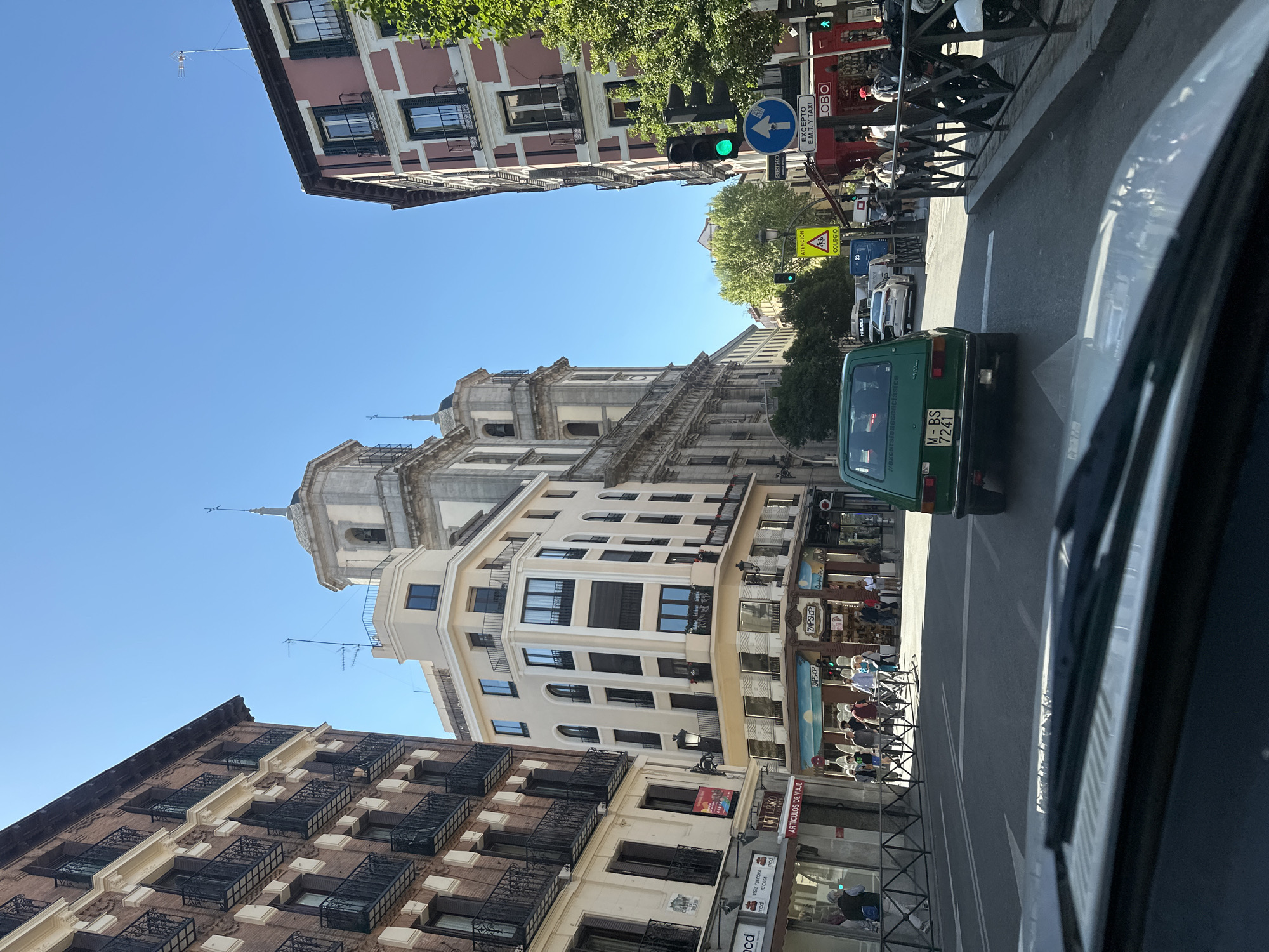 This vibrant street scene, set in Madrid, Spain, captures a bustling daytime environment under a clear blue sky. The image looks down a city street lined with multi-story buildings, featuring both classic European architectural styles with ornate balconies and more modern facades. In the background, a grand, light-colored building with a distinctive dome or bell tower rises above the surrounding structures.
On the right side of the street, a green classic car, possibly a SEAT 600, with the license plate "M-BS 7241" and the hashtag "#excursionesenclasico" visible on its rear, is driving away from the viewer. Other vehicles and motorcycles are parked or moving further down the road. People can be seen on the sidewalks and seated at outdoor cafe tables, enjoying the good weather. A traffic light showing green indicates active traffic flow.
Notable details include various street signs: a blue circular sign with a white arrow indicating a mandatory direction (likely right turn), accompanied by text "EXCEPTO E.M.T. Y TAXI" (meaning "Except EMT and Taxi"), suggesting a dedicated lane or restriction. Another yellow triangular sign warns "ATENCIÓN COLEGIO" (Attention School). Shopfronts are visible at street level, with awnings and outdoor seating. Visible text on shops includes "ARTICULOS DE VIAJE" (Travel Items), "ZAPSHOP" (likely a shoe shop), "SEIKO," and "LOBO." The strong sunlight casts distinct shadows, indicating a bright, clear day.