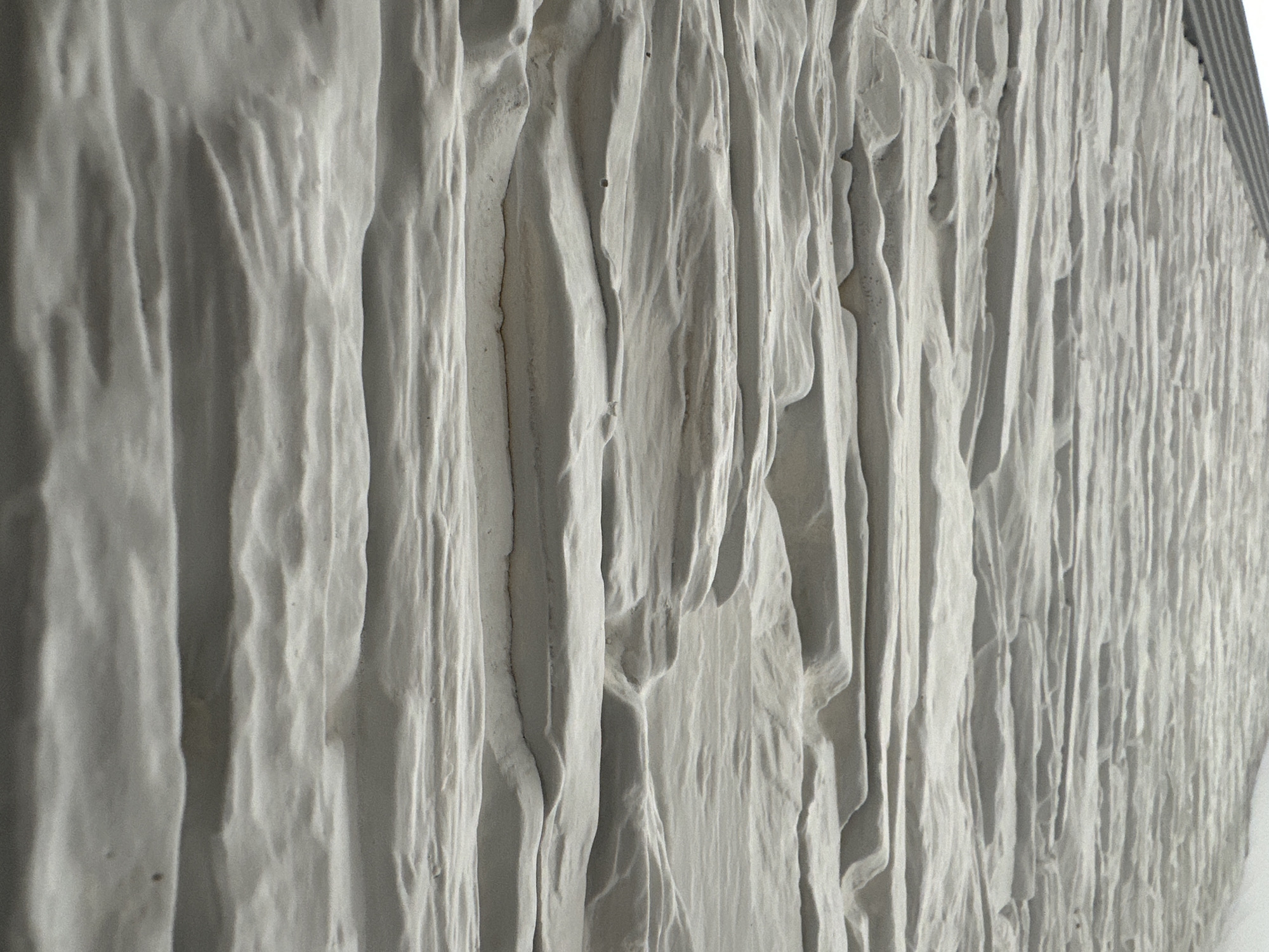 The image presents a close-up, angled view of a highly textured, light-colored surface. The dominant feature is a series of pronounced, undulating vertical ridges and deep grooves, suggesting a wall or a large sculptural art piece. The color palette is predominantly off-white or very light gray, with variations in shade created by directional lighting that casts strong shadows within the crevices and highlights the raised elements.
The texture gives the impression of a solidified flowing material, such as plaster, concrete, or possibly carved stone, or even resembles draped and wrinkled fabric. Its organic, fluid forms create a dynamic three-dimensional effect across the entire frame. The perspective looks along the length of this textured surface, extending from a softer, less defined area on the left to more numerous and slender ridges disappearing into the background on the right.
No individuals or specific objects beyond the textured surface are visible. No activity is taking place; the scene is static and focuses solely on the intricate details of the material and its interaction with light. There are no notable location cues, time of day indicators, or visible text. The image emphasizes abstract form, light, and shadow.